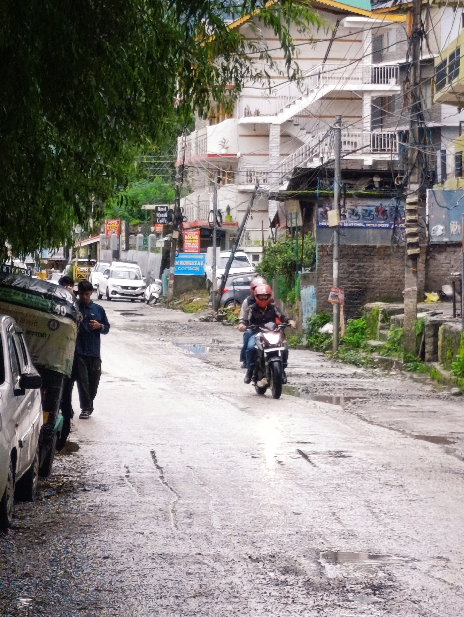 The image depicts a street scene in Chichoga, India, characterized by a wet, uneven asphalt road with numerous potholes and puddles, indicative of recent rainfall. Lush green foliage overhangs from the left side of the frame, partially obscuring a multi-story building with white balconies and an external staircase. The building appears to be a hotel or guesthouse, with signs for "Hotel Mandali" and "Homestay" visible.
In the foreground, a person wearing a dark blue shirt and dark pants walks on the left side of the road. To their left, a portion of a silver vehicle is visible. Further down the street, a motorcycle carrying two individuals wearing helmets rides away from the viewer. The motorcycle rider is wearing a dark jacket and blue jeans, and the pillion rider is wearing a lighter colored jacket. Various vehicles, including a white SUV and a white scooter, are parked or driving on the street in the mid-ground. The overall atmosphere suggests a busy, somewhat chaotic urban environment, possibly a tourist area, with visible electrical wires and poles adding to the density of the scene. The weather appears overcast, contributing to the wet conditions.