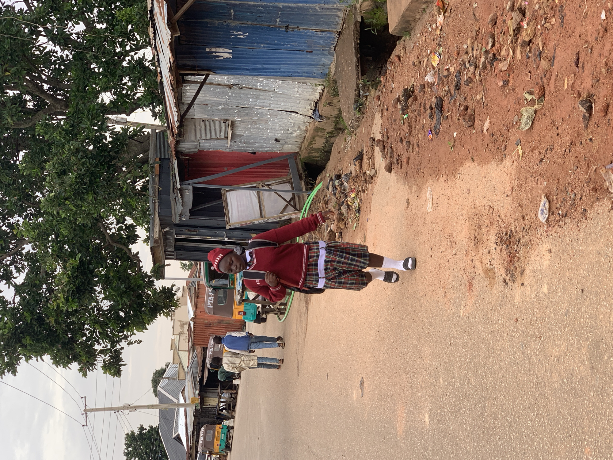 An overhead shot shows a girl in a school uniform walking on a dusty road in the city of Jos, Nigeria. The girl is wearing a red sweater over a white collared shirt, a plaid skirt, white socks, and black sandals. She has a backpack slung over her right shoulder. The road is lined with makeshift shops made of corrugated metal. In the background, there are several three-wheeled vehicles, commonly known as "Keke" or "Marwa," and a utility pole with wires. The ground is covered with red dirt and scattered debris, suggesting a somewhat unkempt environment. A large tree with green leaves overhangs the scene from the left. The sky is overcast.