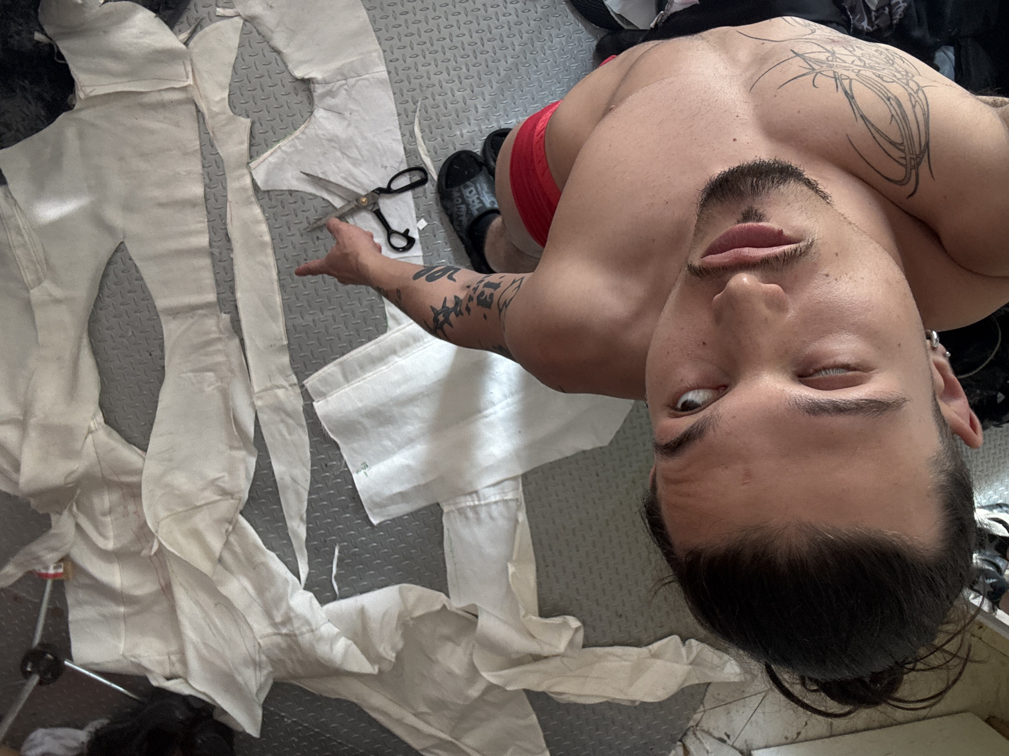 A young male with tattoos on his arm and shoulder is lying on a textured grey floor, looking up at the camera. He has dark hair pulled back in a bun, a goatee, and is wearing red shorts and black slides. The floor is covered with pieces of white fabric, some of which appear to be cut from a garment. A pair of scissors is on the fabric near his right hand. The scene appears to be indoors, potentially a workshop or studio. There is no indication of the city or country.