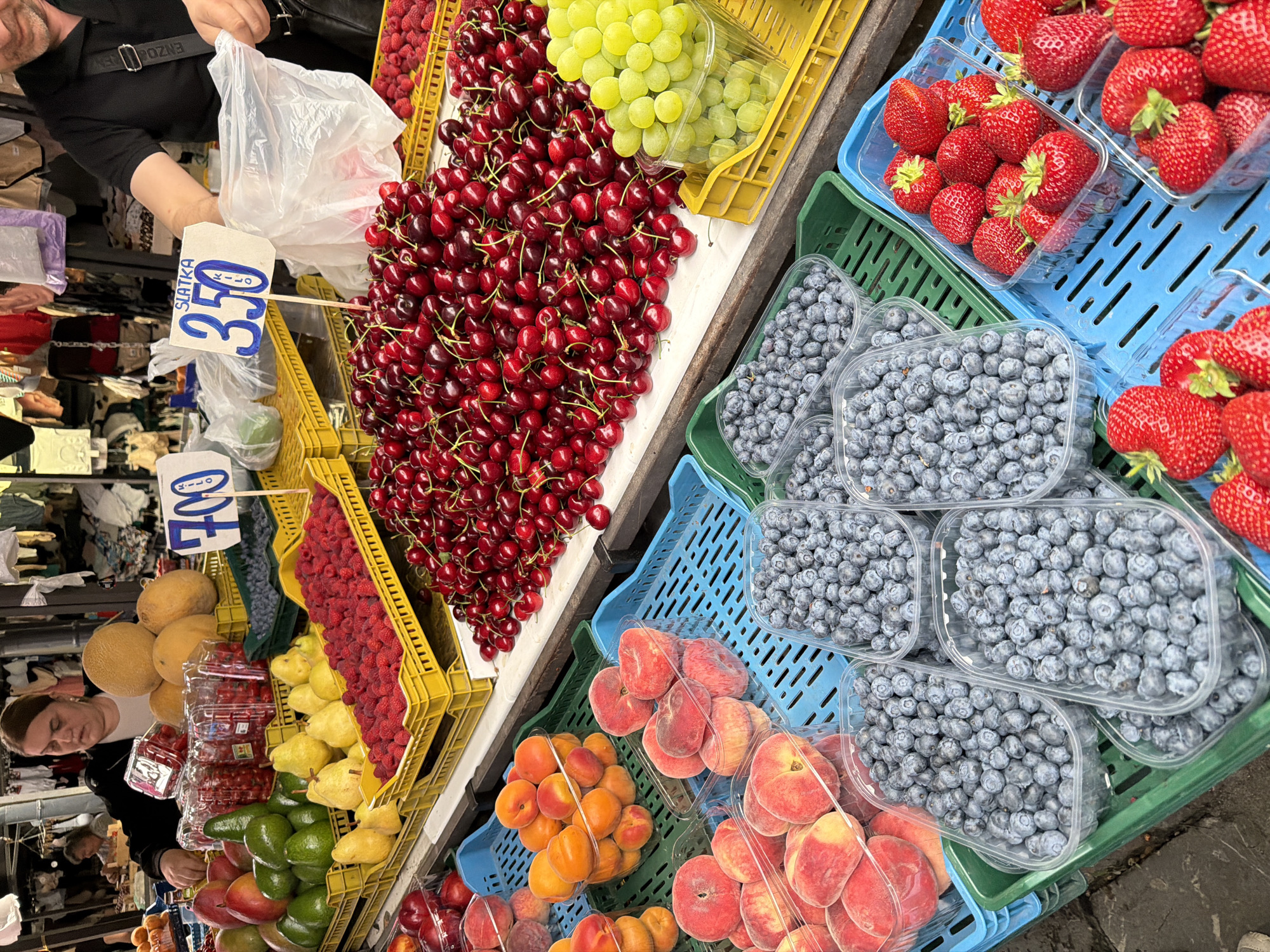 A bustling outdoor market stall in Vračar Urban Municipality, Serbia, displays an abundant selection of fresh fruits. In the foreground, various containers overflow with vibrant strawberries, their red hues contrasting with the deep blue of blueberries. To the right, a crate of bright green grapes adds another pop of color. Behind these, large piles of ripe cherries are showcased, with a smaller assortment of apricots visible further back. The stall also features rows of peaches and yellow pears, alongside avocados and what appear to be red currants in plastic containers. Melons are stacked on top, and a vendor, a woman with dark hair wearing a dark jacket over a white shirt, is seen arranging the fruit. Another vendor, a man with dark hair and a black shirt, is visible on the right, handling a plastic bag. Price signs, one reading "700 KILO" and another "SLATKA 350 KILO," are displayed, indicating the market's commerce. The scene is captured under natural lighting, highlighting the freshness and variety of the produce.