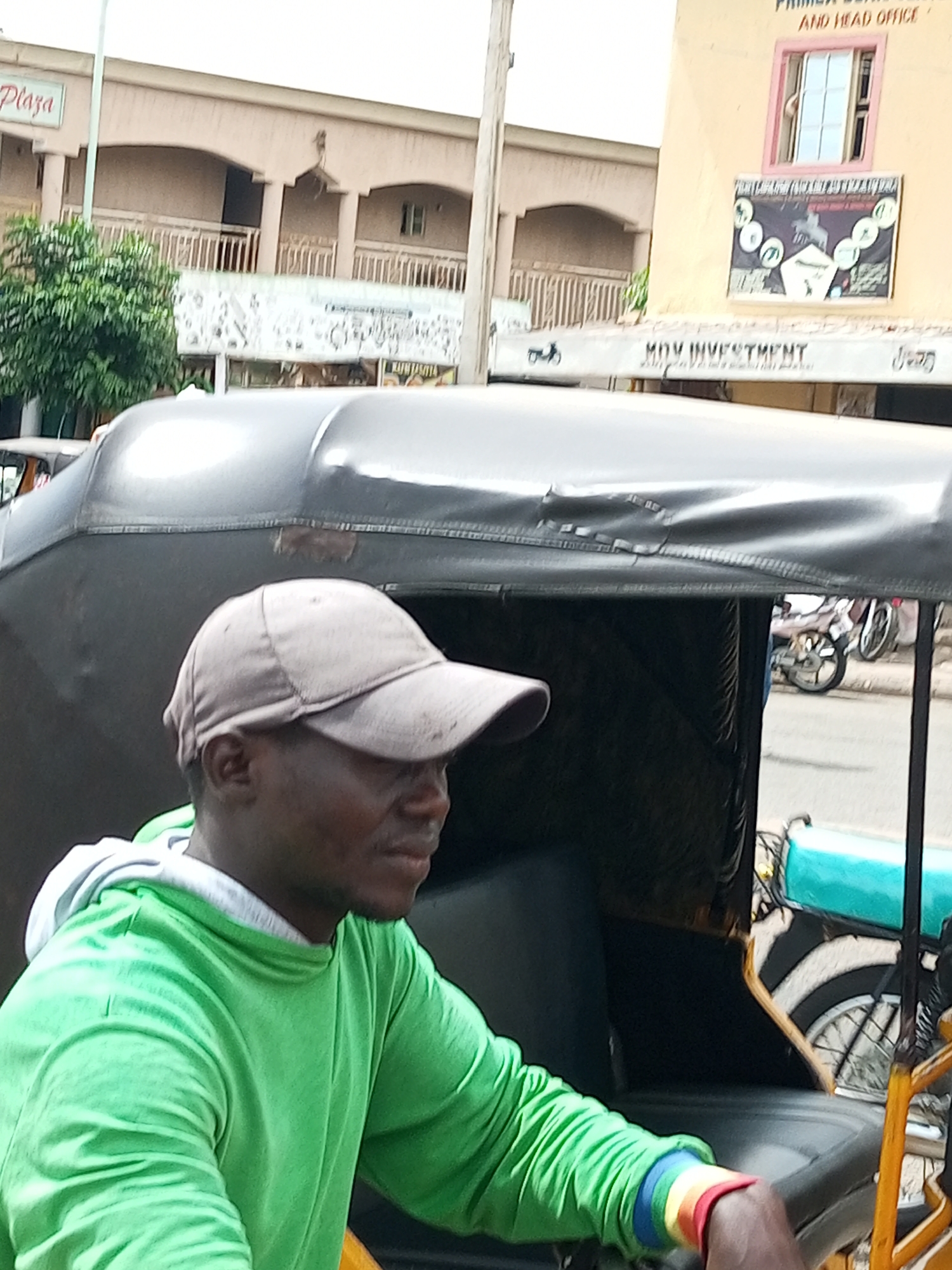 The image shows a close-up view of a man wearing a light brown baseball cap and a bright green hooded sweatshirt. He is sitting in what appears to be a tricycle taxi, commonly known as an "okada" or "keke napep" in Nigeria. The man's face is partially visible, and he is looking to his right. His left arm, adorned with a wristband featuring rainbow-colored stripes, is resting on the side of the vehicle.
In the background, a building with arched walkways and balconies suggests an urban setting. A sign with the word "Plaza" is visible on the upper left side of the building. To the right, another building with a yellow facade displays signage, including a banner with graphics and text, and another sign reading "MDV INVESTMENT". A window on this building shows a person's hand. The street is visible in the lower right portion of the image, with a motorcycle parked on the side. The overall scene captures a slice of everyday life in a Nigerian city, likely Arawa, given the context.