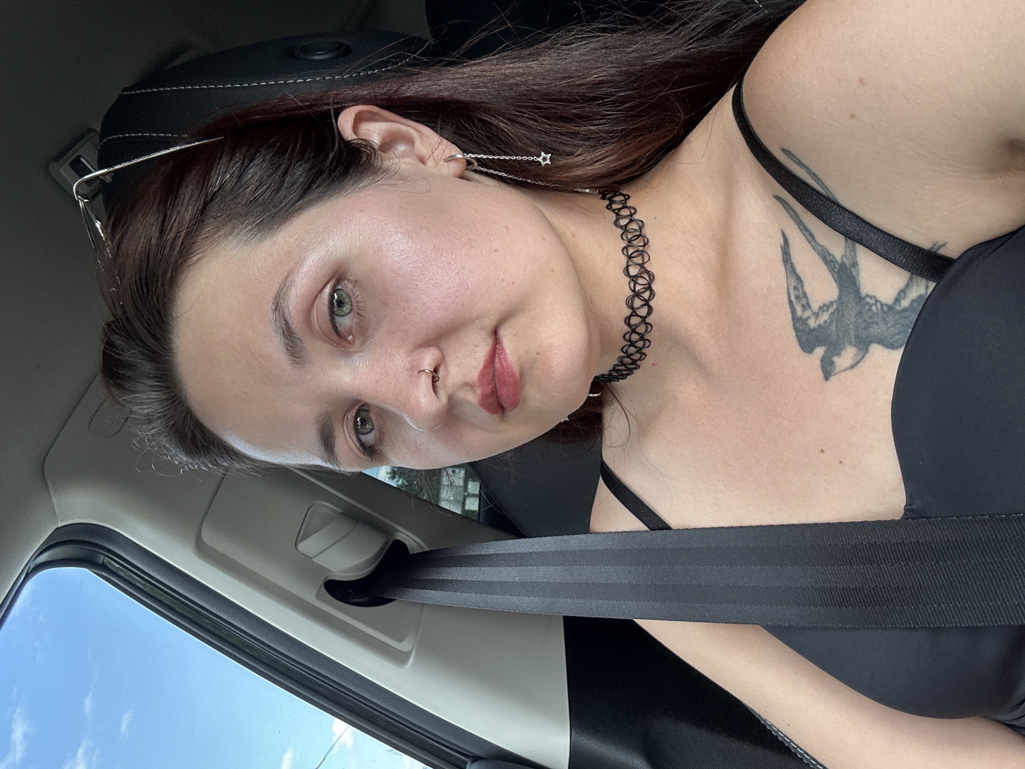 A young woman with long, dark hair and green eyes is shown from the chest up, sitting in the passenger seat of a car. She is wearing a black bra, a black choker necklace, and a long silver earring with a star charm. A black seatbelt is fastened across her chest. A swallow tattoo is visible on her left shoulder. The background shows a blue sky with white clouds and the interior of the car, including a headrest and a window. The context of Naberezhnye Chelny, Russia is not discernible from the image itself.