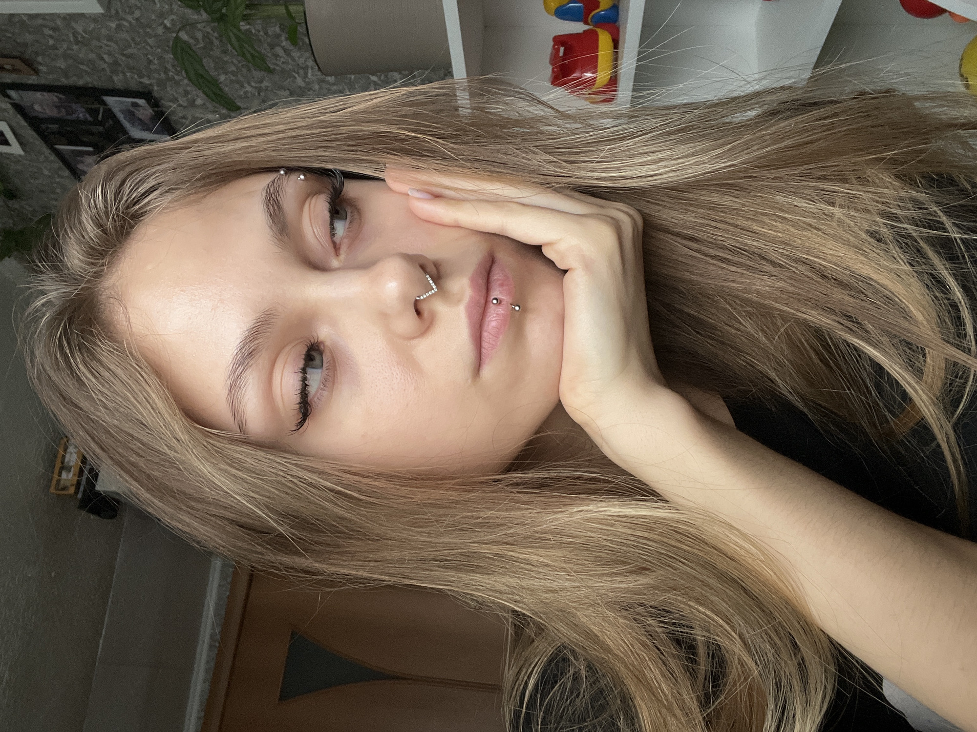 A blonde woman is posing for a selfie. She has several piercings: one in her eyebrow, one in her nose, and one below her bottom lip. She is holding her face with her left hand. The background is a room with a shelf and a plant. It is not possible to determine the location in the city of Фоки, Russia from the given image.