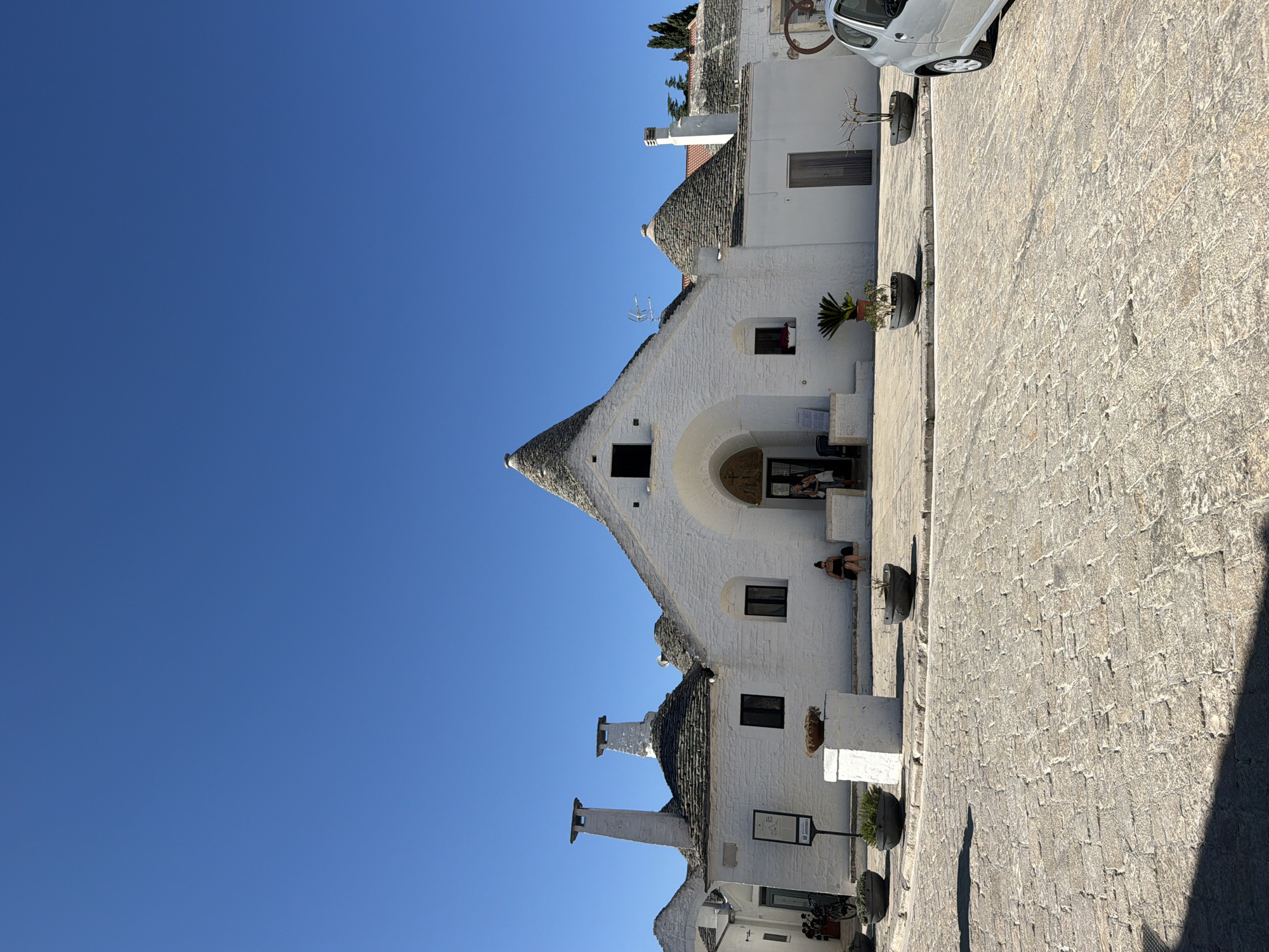The image depicts a sunny daytime scene in Alberobello, Italy, showcasing the iconic Trulli houses. In the foreground, a cobblestone street is visible, with shadows cast by unseen objects. Several Trulli houses, characterized by their conical roofs and whitewashed walls, line the street. Some of these structures have chimneys protruding from their roofs. Planters with small green plants are placed near the houses, and one planter sits atop a white pedestal. Two women are seated on a step in front of one of the Trulli houses. To the right, the rear of a silver car is partially visible. The sky is clear and blue.