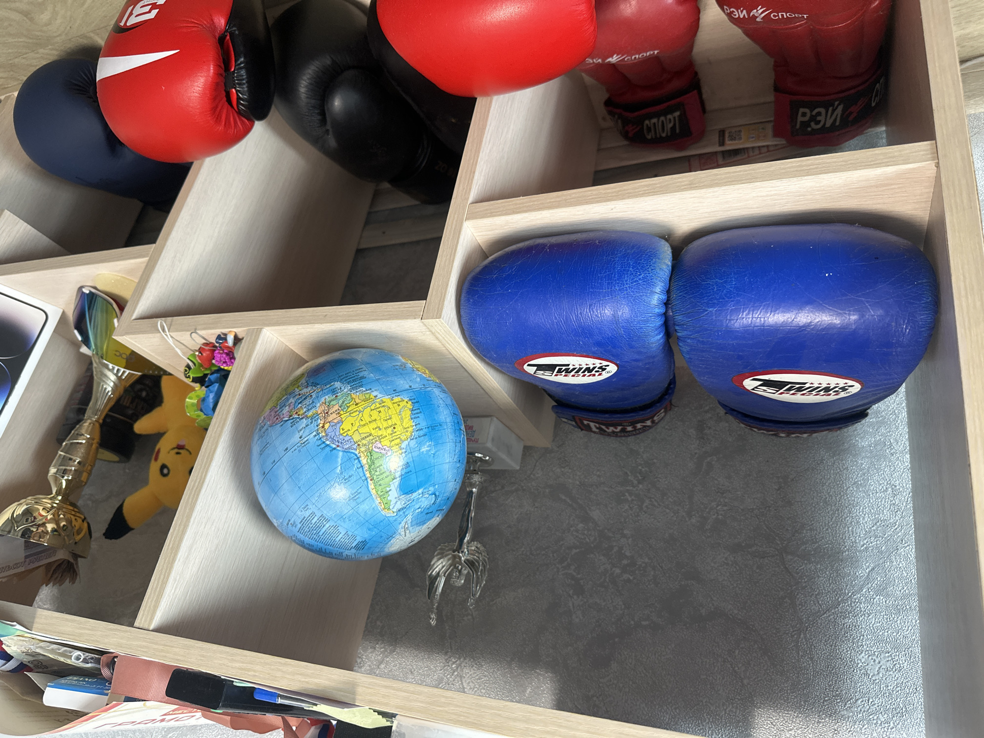 This is a still, eye-level, medium shot of boxing gloves and other items on a wooden shelf. The shelf is light-colored wood with cubbies. The background is a textured gray wall. In the top cubby, there are two blue boxing gloves with white lettering. To the right of the gloves is a small globe. To the right of the globe is a small statuette of a winged figure. In the cubby below, there are two red boxing gloves with white lettering. To the right of these are black boxing gloves and another blue boxing glove. A red boxing glove is in the foreground. The scene is indoors and appears to be a storage area or display shelf. There are no people visible in the image. The location is described as being in the city of Birobidzhan, Russia, but there is no visual information in the image to confirm this.