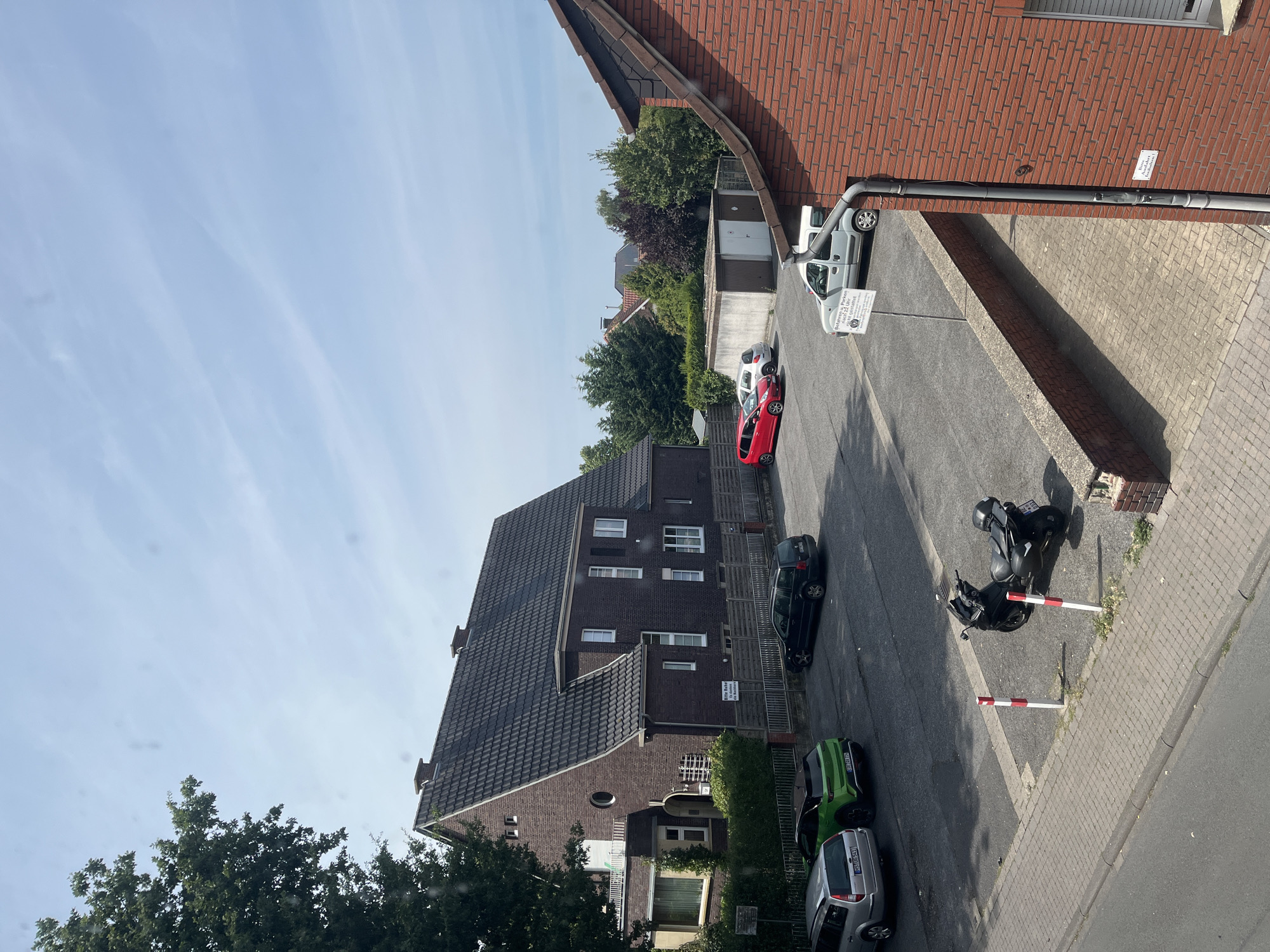 The image captures a street scene in Ascheberg, Germany, on a clear, sunny day. In the foreground, a red brick building with a sloped roof is visible on the right. Adjacent to it is a paved area with a low stone wall and a sidewalk. Across the street, a dark asphalt parking lot is occupied by several vehicles. These include a silver station wagon, a bright green hatchback, a black station wagon, a red compact car, a white compact car, and a light gray minivan. A black scooter is parked in the lot. In the background, a large, dark-tiled roof house with multiple windows is prominent, flanked by trees and other buildings. The sky is blue with scattered white clouds.