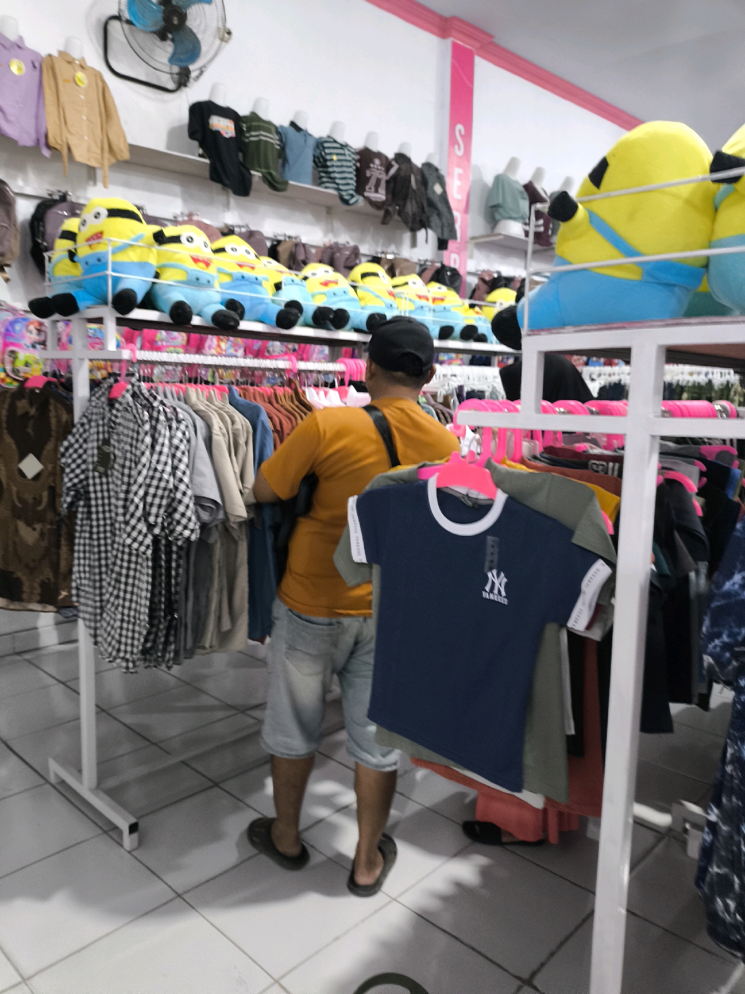 This image depicts the interior of a retail store, likely a clothing boutique, located in the city of Posso, Indonesia. The scene is brightly lit with fluorescent lighting reflecting off the tiled floor. In the foreground, a man wearing an orange t-shirt and denim shorts is standing with his back to the camera, facing a display rack of clothing. He appears to be browsing or examining the merchandise. Several racks of clothes are visible throughout the store, including shirts, jackets, and plush toys, some of which are shaped like characters from the movie Despicable Me. The shelves are stocked with a variety of clothing items, many of which are displayed on mannequins. The overall impression is that of a busy, well-stocked store.
