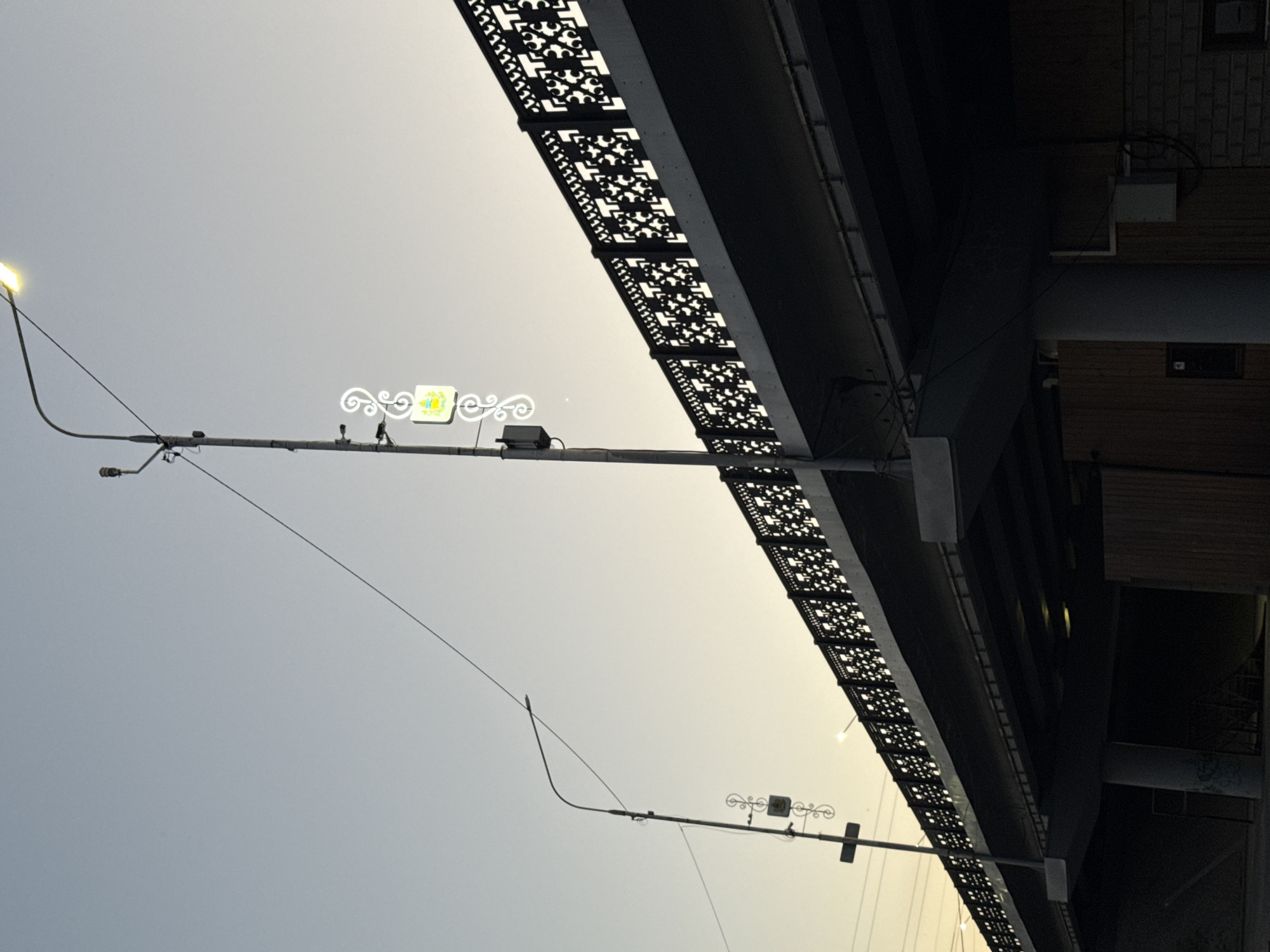 This is a media file, likely a photograph, showing a view looking up towards an elevated structure, possibly a bridge or overpass, against a pale, overcast sky. The perspective is tilted, making it appear as if the scene is sideways.
The main subjects are structural elements. A prominent object is a streetlight with a single light fixture, casting a warm, yellow glow. Extending from this streetlight is a horizontal arm supporting a decorative element with a yellow and white illuminated sign. The sign has a stylized floral or crest-like design. Wires are visible, strung between various points on the structure and the streetlight.
Underneath the elevated structure, decorative panels with intricate, dark cut-out patterns are visible. These panels are affixed to the underside of the structure, creating a rhythmic visual effect. Further to the right, parts of a building facade are visible, constructed with wooden planks and brickwork. A small, dark rectangular opening, possibly a window, is set into the wooden section.
The time of day appears to be late afternoon or early evening, suggested by the soft, diffused light and the illuminated signs. The weather seems to be cloudy or hazy, contributing to the muted tones of the sky. There are no people clearly visible. The overall mood is calm and somewhat industrial due to the visible infrastructure. No discernible text is present within the image. The location context provided is Novyi Chirkei, Russia.