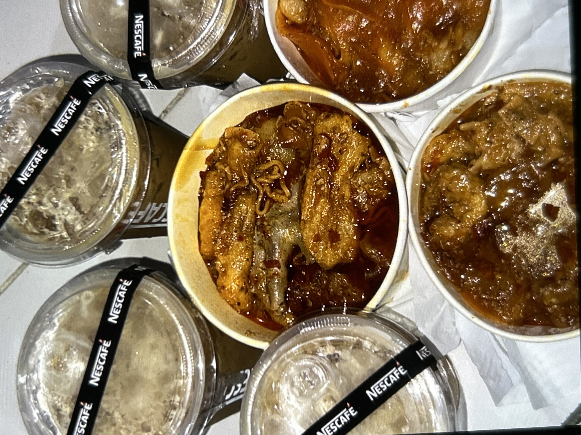 A top-down, close-up shot captures four disposable cups of Nescafe coffee and three paper containers of food. The coffee cups, some sealed with plastic lids, are arranged around the food containers. A black band with the Nescafe logo encircles each coffee cup. The food containers are filled with what appears to be braised or stewed dishes in rich, dark sauces. One container in the center holds pieces of meat or tofu and what seems to be noodles. The other two containers contain similar saucy dishes. The items are placed on a white, textured surface, possibly a table or a tray, with some white paper napkins visible. The lighting is soft and diffused, creating a slight sheen on the food's sauces. The scene suggests a casual takeout meal. The image does not contain any identifiable landmarks or indicators of its specific location in Depok, Indonesia.