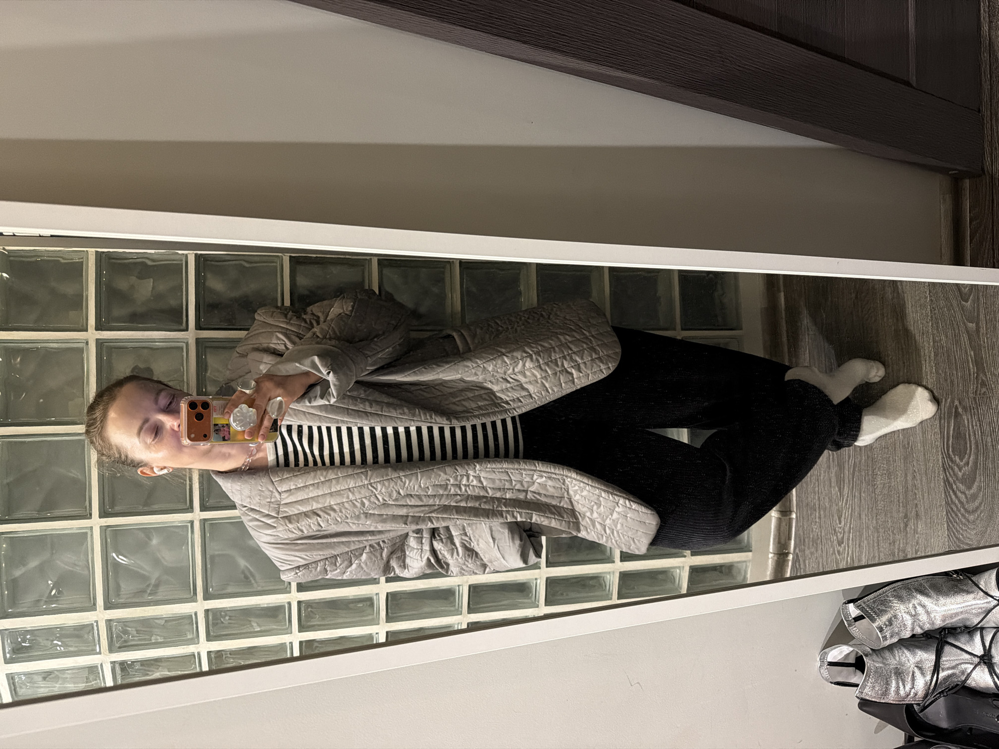 A person is reflected in a mirror. The person is wearing a gray quilted jacket over a black and white striped shirt, and black pants with white socks. They are holding a phone up to their face. The reflection also shows a wall of glass blocks, a gray wall, a dark wood molding, wooden flooring, and a silver-colored object on the floor. There is no information available to conclude that the image was taken in Moscow, Russia.
