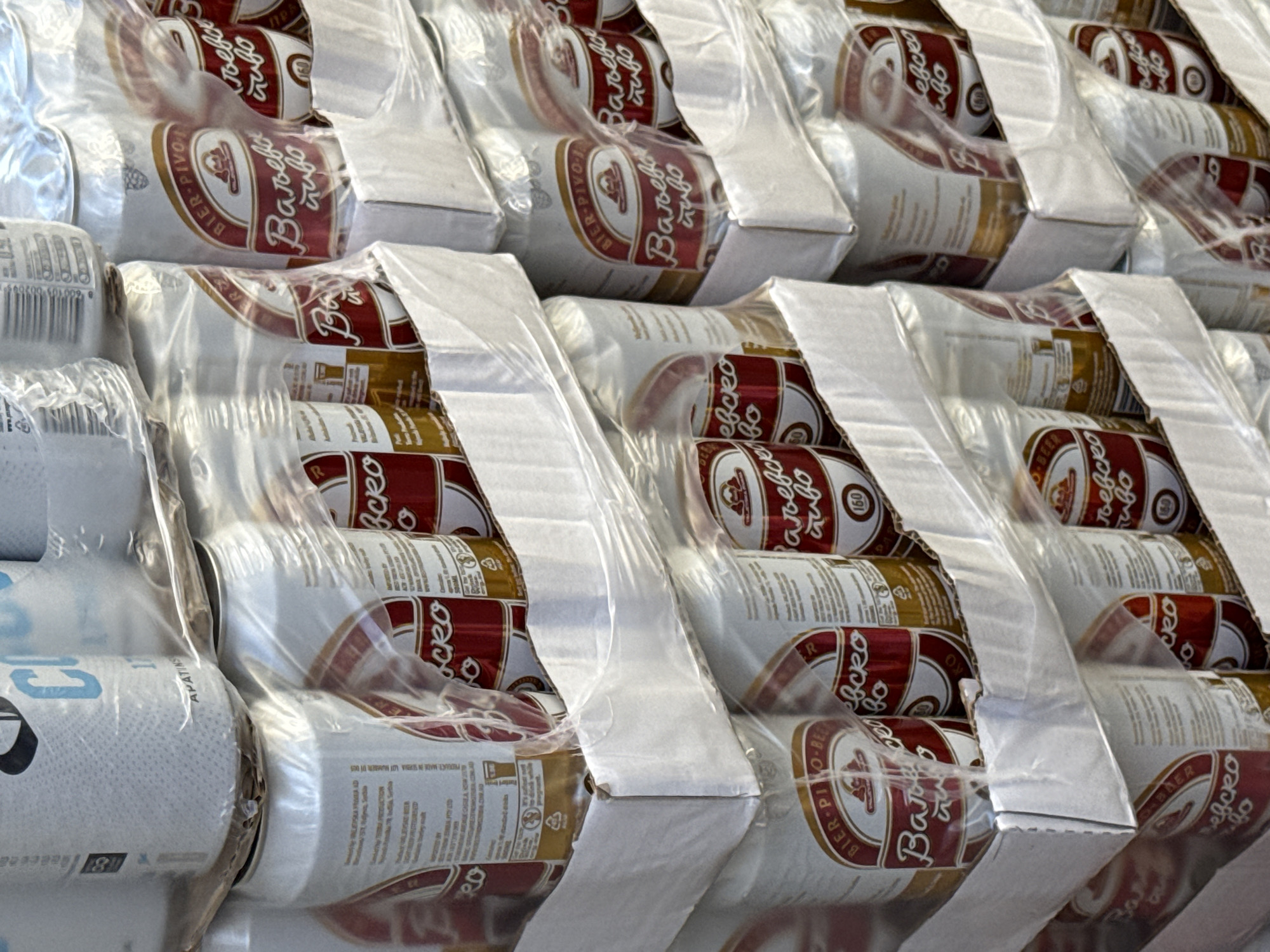 The image depicts a close-up of several stacked six-packs of canned beer, possibly in a retail environment or storage area. The beer brand appears to be "Valjevsko pivo," a Serbian beer. The cans are shrink-wrapped in plastic. One can in the foreground is partially visible and marked with the word "APATINSKO". There are details of the brewery printed on the side of a can stating "PRODUCT MADE IN SERBIA LOT NUMBER: 01 055". The location of the image is not evident from the visual data, but as the beer is from Serbia, it is possible the location is in the city of Belgrade. There are no people visible in the image. The scene appears to be a common commercial setting of stacked and packaged goods.