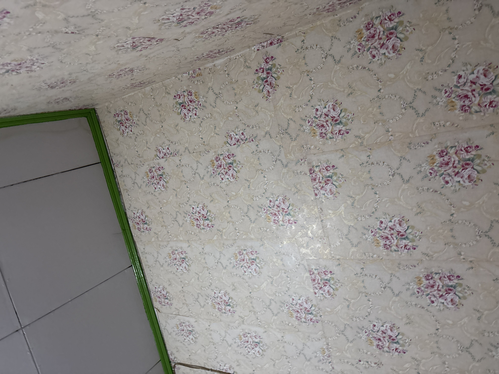 The image shows a close-up, high-angle shot of a wall covered in floral wallpaper. The wallpaper is a light beige color with repeating patterns of pink and white roses, accented by delicate green leaves and swirling vine-like designs. The wallpaper appears to be applied to a wall that is meeting another wall at a corner, suggesting an interior room. A bright green trim runs along the top edge of the wallpaper where it meets the ceiling. The ceiling itself is visible in the upper left quadrant of the image and appears to be made of white rectangular panels. The overall impression is that of a domestic interior, possibly a bathroom or a bedroom, given the wallpaper pattern and the presence of the green trim and ceiling. The lighting suggests an indoor environment, possibly with artificial light. The specific location in Palimanan, Indonesia, is not discernible from the image itself.