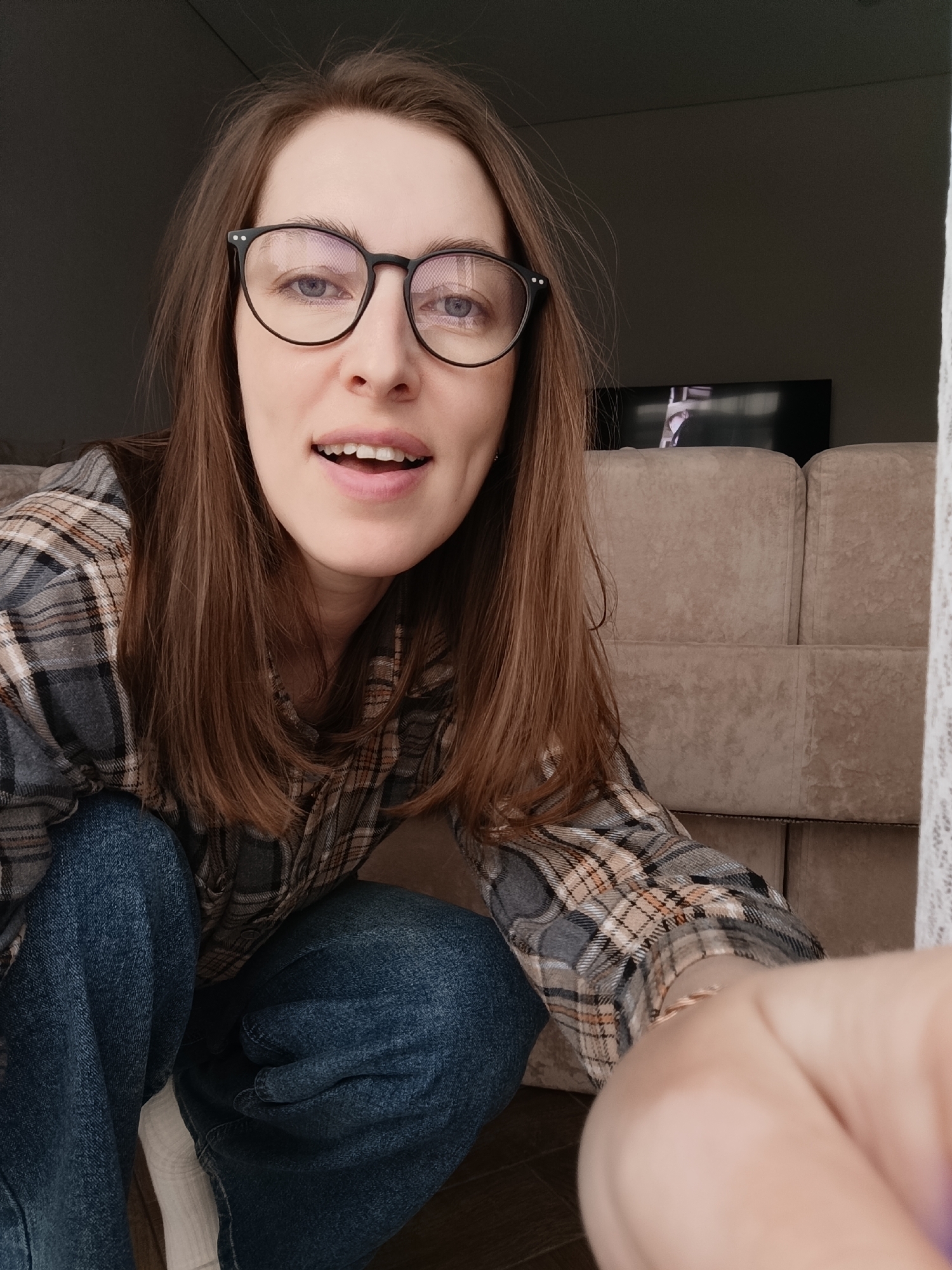 The image depicts a woman with long, straight brown hair and wearing black-rimmed glasses, crouching indoors. She's wearing a plaid shirt in shades of grey, brown, and tan, and blue jeans. A portion of a white sock is visible near her ankle. Behind her is a light-colored, possibly beige, upholstered sofa. In the background, a black television screen is visible. The photo appears to be taken from a low angle, suggesting it might be a selfie. The location is indicated as Chaikovsky, Russia, though this is not visually discernible from the image itself.