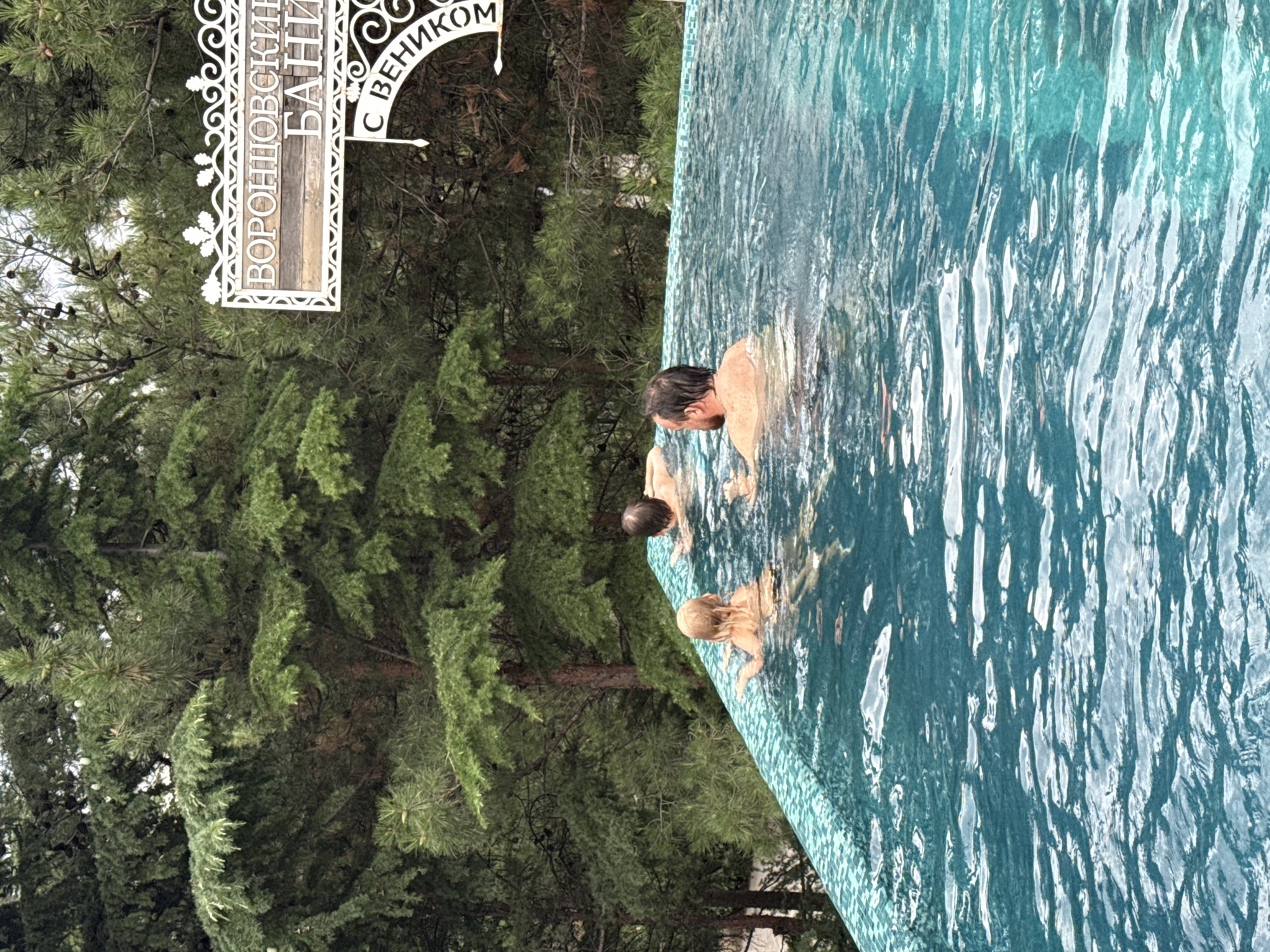 In the city of Yalta, Ukraine, a refreshing scene unfolds in an infinity pool nestled amidst a lush backdrop of evergreen trees. The vibrant turquoise water shimmers, reflecting the dappled sunlight filtering through the dense foliage. Three individuals are present in the pool. A man with a receding hairline and a beard is seen from the back, his arms submerged in the water. To his left, a child with blonde hair is partially visible, also in the water. Further to the left, another child's head and shoulders can be seen above the water's surface. A white ornate sign, featuring Cyrillic lettering that appears to read "ВОРОНЦОВСКИЕ БАНИ", is prominently displayed against the green canopy of the trees, suggesting a destination associated with baths or saunas. The overall ambiance is one of relaxation and enjoyment in a natural, serene setting.