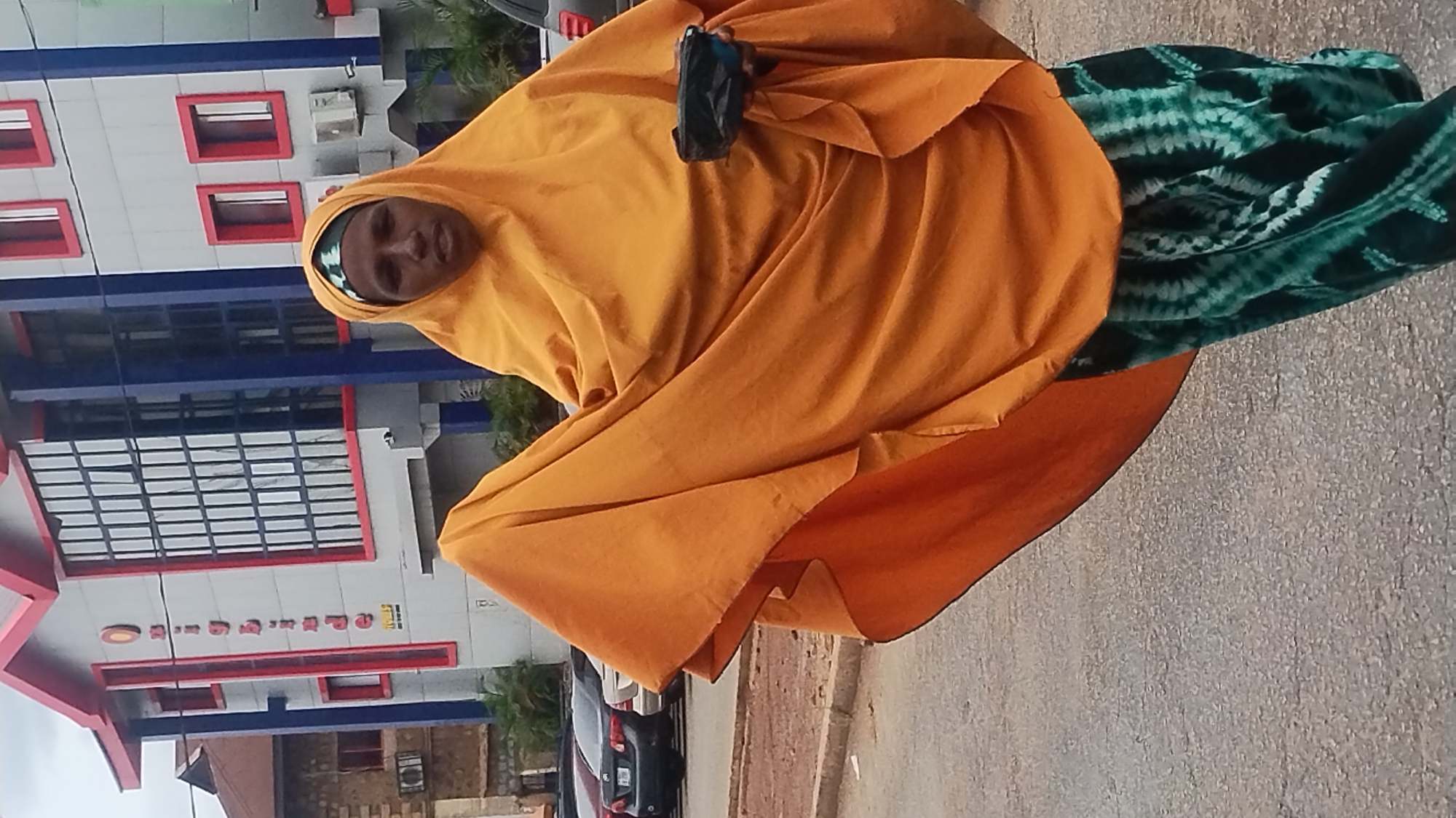 The image captures an individual standing outdoors in what appears to be a street setting in Jos, Nigeria, during daylight hours. The person, with dark skin, is adorned in a flowing, bright yellow or orange head covering (likely a hijab or khimar) that covers most of their body. A distinct green and white patterned garment is visible beneath the outer covering, particularly on the left side of the frame. In their right hand, the individual holds a small, dark object, possibly a mobile phone or a compact bag.
The background features a multi-story building with a modern design, characterized by a light-colored facade with prominent blue and red architectural accents. Multiple windows are visible across the building's surface, some framed in red, and air conditioning units are mounted on its exterior. The building also displays sections with glass panels, reflecting the environment. Below the building and to the right of the person, a paved street or sidewalk is visible. Several cars are partially visible further in the background, suggesting a bustling urban or semi-urban environment. The lighting indicates it is daytime, possibly bright or slightly overcast, as no harsh shadows are cast. The person's facial expression is neutral and not clearly discernible due to the angle and partial obscuration. Some faint, illegible text appears on a red band of the building's facade.