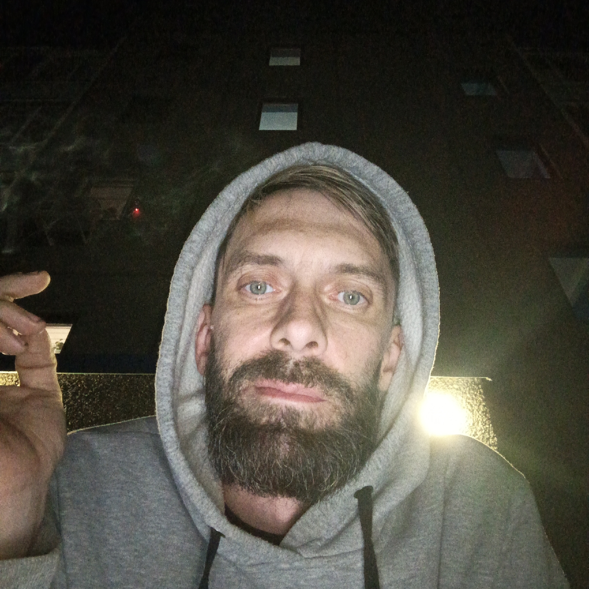 A night-time selfie captures a man in Марковский, Russia. He looks directly at the camera with a neutral expression, revealing bright blue-green eyes, a full beard, and a mustache. He is wearing a grey hooded sweatshirt with the hood pulled up. His left hand is partially visible in the upper left corner of the frame, with fingers slightly raised.
The background is very dark, indicating it is night. Above his head, faint, dark rectangular shapes, likely windows of a building, are visible against the dark sky; some show faint reflections. To the right of the man, a strong, bright light source creates a prominent flare and illuminates his face from below and slightly to the side. A subtle red light dot can also be seen in the distant upper-left background, possibly an indicator light. There is no visible text in the image.