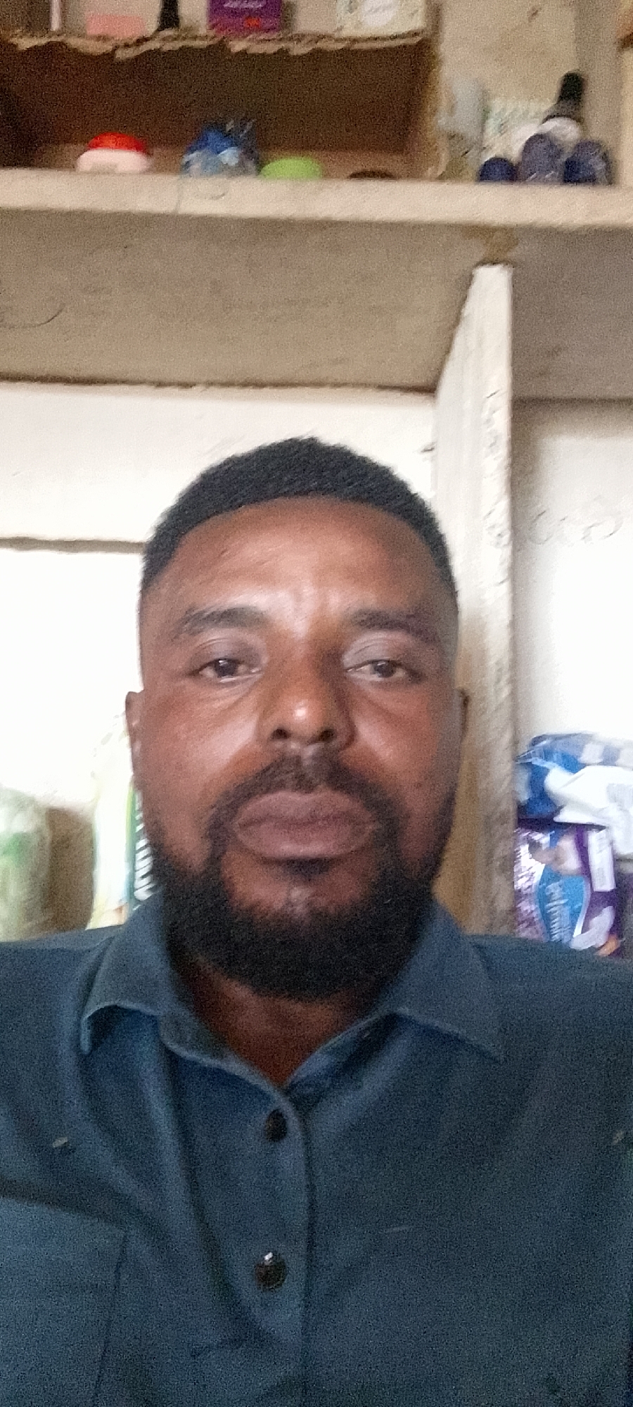 The image shows a medium close-up of a man with dark skin, a dark beard, and short, dark hair, wearing a dark blue collared shirt. He is looking directly at the camera. The background appears to be an indoor setting with shelves holding various objects, including what look like packaged goods and possibly some containers. The lighting is moderate, and the image quality is fair. The specific location within Durumi, Nigeria is not discernible from the visual information provided.