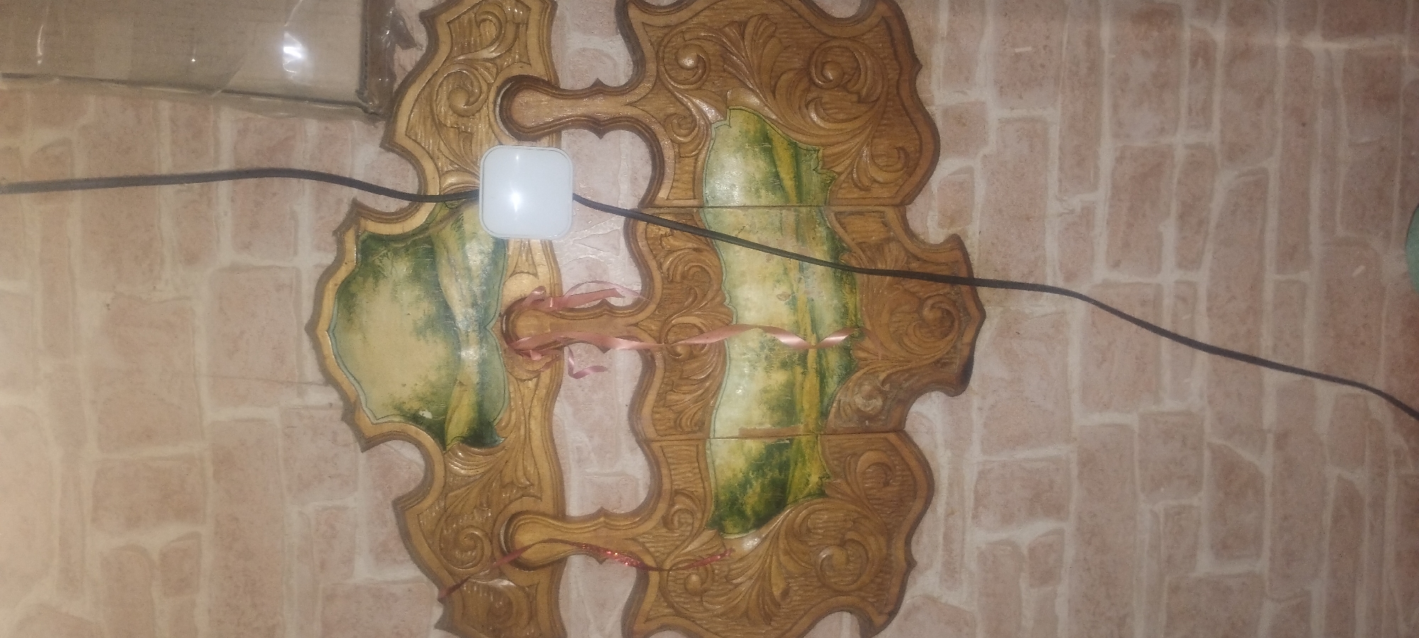 The image shows a close-up view of a decorative wall hanging with a landscape painting, possibly in an interior space with a brick-patterned wall. A black electrical cord runs vertically down the center of the image, passing over the wall hanging and a small white square object, likely a device or fixture. There are pink ribbons attached to the wall hanging. The brick pattern of the wall is light brown and appears to be made of faux bricks. There is a clear plastic covering visible on the upper right side of the image. The lighting suggests the image was taken indoors, possibly at night or in a dimly lit area.