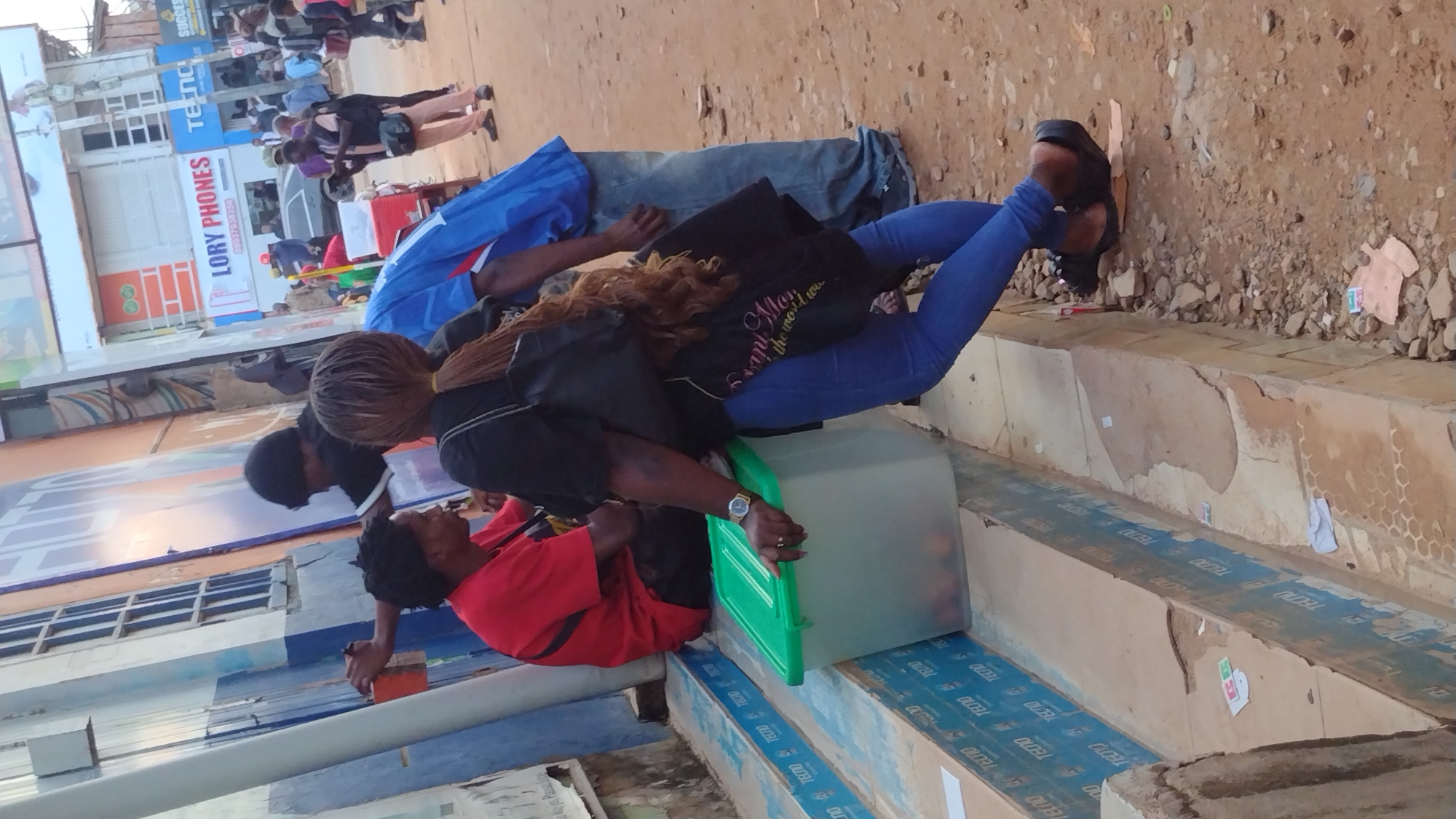 The image depicts a street scene in Jos, Nigeria. In the foreground, a woman wearing blue jeans and black sandals leans against a green plastic container. She has long, braided brown hair and a black bag slung over her shoulder. Next to her, a man in a red shirt is seated. Behind them, another person, partially obscured, wears a black beanie and a black and white jacket.
The environment includes a building with blue and orange signage, featuring text like "LORY PHONES". The ground is unpaved and covered with dirt and scattered debris. In the background, other individuals are visible, some appearing to be patrons or employees of the businesses. The overall scene suggests a busy commercial area.