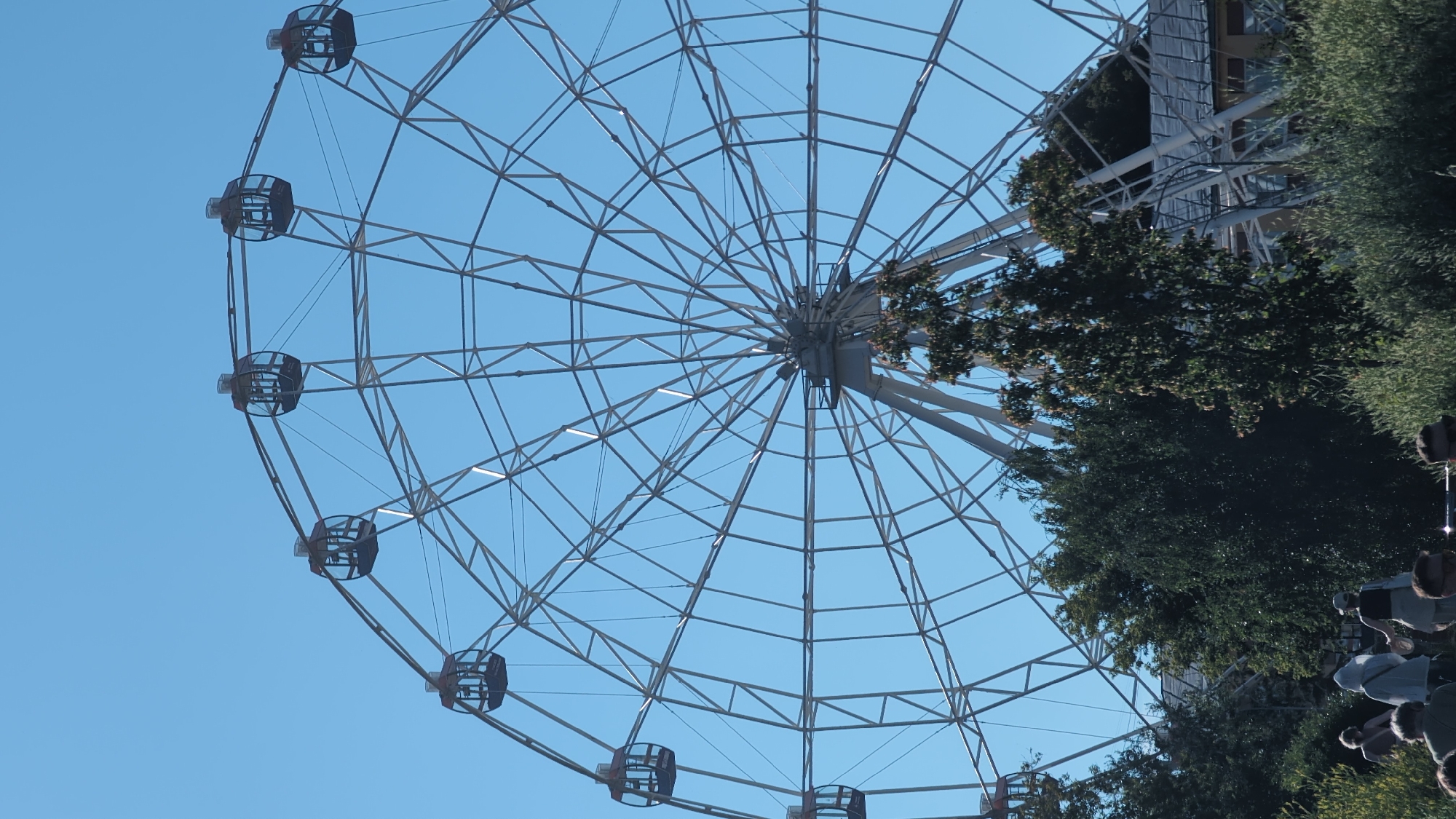 A large, silver Ferris wheel dominates the frame against a clear blue sky. The wheel has a complex metal structure with numerous spokes radiating from the center. Several enclosed, dark-colored gondolas are visible, attached to the outer rim of the wheel. In the foreground, the tops of several trees with green foliage are visible, partially obscuring the lower part of the Ferris wheel structure and some buildings. A few people are present in the lower part of the image, walking among the trees and greenery. The scene appears to be outdoors, likely in a park or amusement area. This Ferris wheel is likely the "Molodezhnaya" Ferris wheel, a prominent landmark in Nizhny Novgorod, Russia, situated in the Molodezhnaya Park.