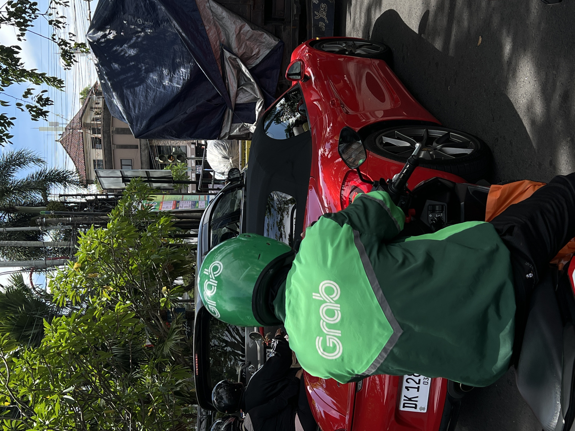 The image captures a bright, sunny street scene in Denpasar, Indonesia, showing a Grab delivery rider on a motorcycle from a rear perspective. The rider wears a distinctive green helmet and jacket with the "Grab" logo prominently displayed in white. The rider is stationary, possibly in traffic, positioned behind a sleek red sports car.
The red car, possibly a Porsche, is also stationary. Its rear license plate, "DK 128...", is clearly visible, with "DK" confirming its registration in Bali. To the left of the red car, another dark-colored vehicle is partially visible, and further down the street, another individual on a motorcycle wearing a black helmet is seen.
In the background, a multi-story building with a traditional tiled roof is visible under a clear, bright sky. Numerous power lines crisscross the sky. Lush green trees and foliage line the street, casting strong shadows on the asphalt, indicating midday or early afternoon. A large blue tarp covers what appears to be a structure or vehicle to the left of the red car. Partial views of traffic lights and other street signage are also present. The overall scene depicts a typical busy urban environment in Southeast Asia.