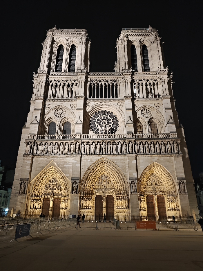 The image captures the illuminated facade of Notre Dame Cathedral in Paris, France, at night. The grand Gothic structure stands against a dark, clear sky, with its intricate details highlighted by artificial lighting.
The cathedral's west front features two prominent rectangular towers at the top. Below the towers, a large rose window with delicate tracery and sculptural figures is visible in the central section. Smaller arched windows flank the rose window. A continuous band of statues, known as the Gallery of Kings, spans the facade above the main portals.
At ground level, three deeply recessed and highly ornamented arched portals serve as the main entrances, each intricately carved with religious scenes and figures.
In the foreground, metal crowd control barriers cordon off the area directly in front of the cathedral. A few individuals are discernible near these barriers and the entrances, appearing as distant silhouettes, possibly pedestrians or visitors.
The presence of the barriers and visible signage indicates restricted access, suggesting ongoing work or temporary closure. An orange sign on a barrier clearly reads "Fermeture temporaire" (Temporary closure) and partially "SR Drive". Another blue sign with a white arrow is also visible on the left side. The scene conveys the majestic presence of the historic landmark under the night sky.