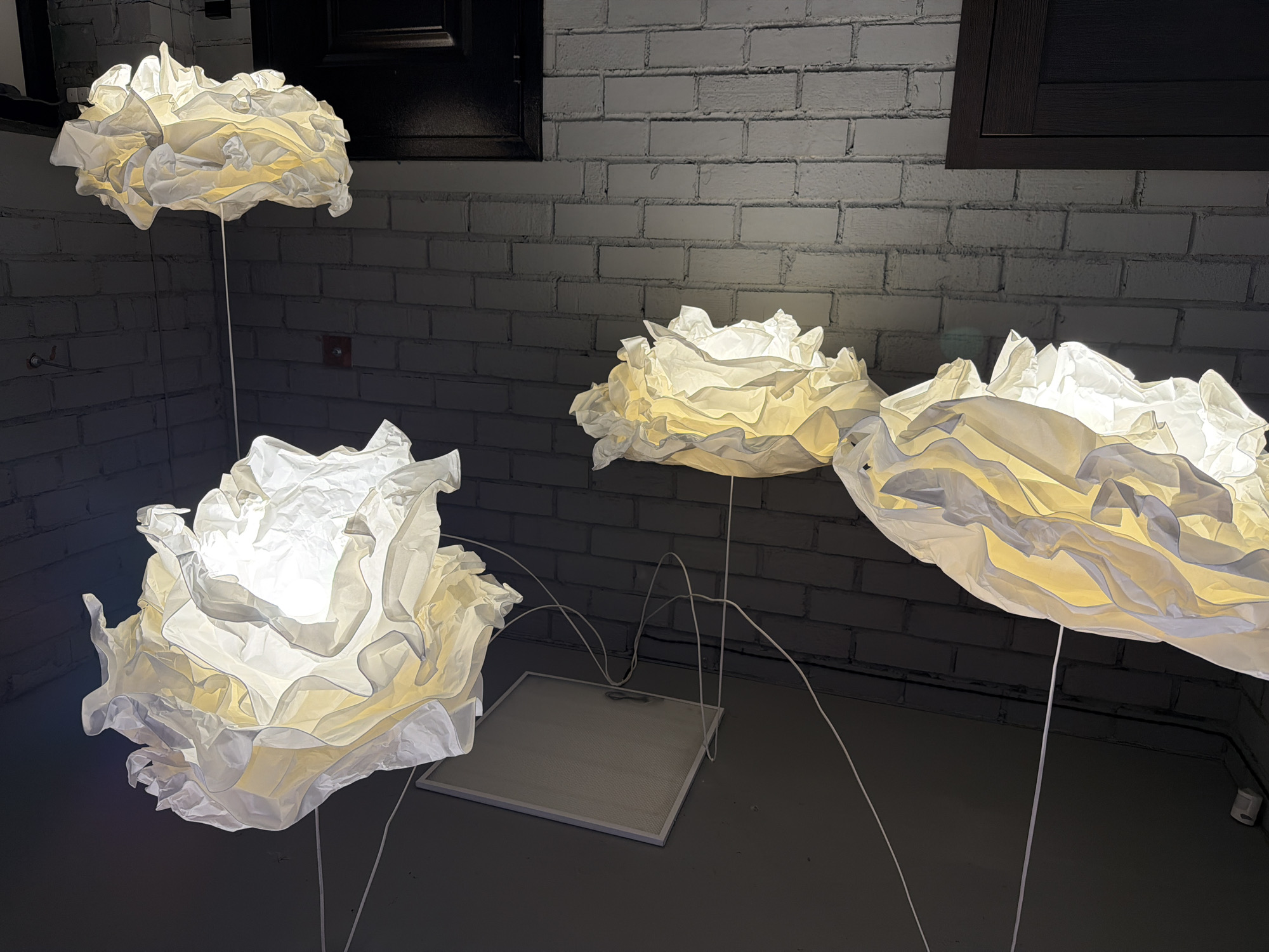 The image displays a dimly lit interior scene with a white brick wall in the background. Three lamps with ruffled, translucent shades, resembling clouds or flowers, are suspended from the ceiling by white cords. The lamps emit a warm, yellow-white light, illuminating the textured brickwork. One of the lamps is larger and positioned to the right, while two smaller ones are to the left. A square ceiling panel is visible above and between the lamps. The overall aesthetic suggests a modern, possibly retail or restaurant setting. There are no people or animals present in the image, and no specific location in Pavlodar, Kazakhstan, can be identified from the visual evidence provided.