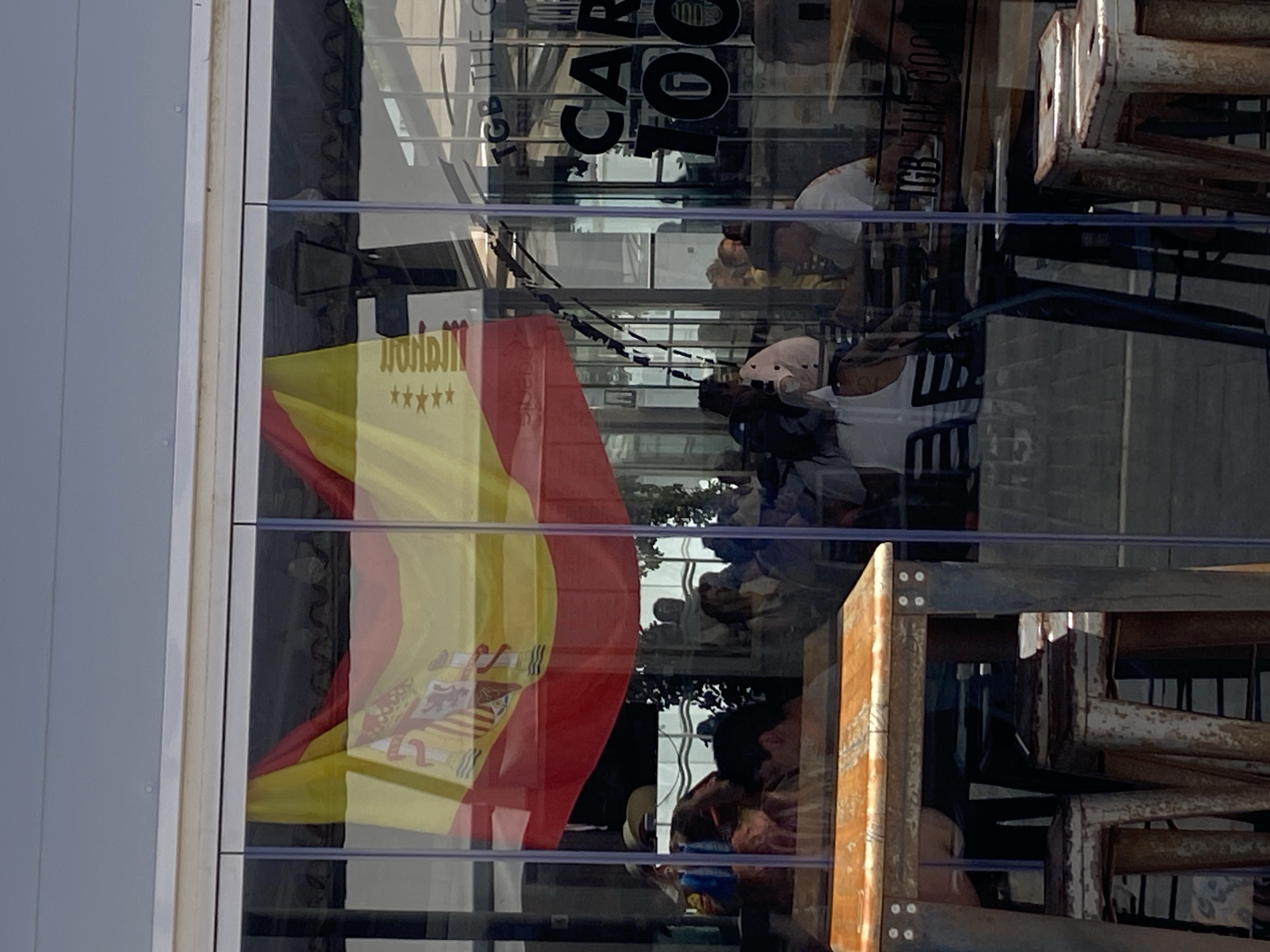 The image captures a view through a glass-fronted establishment, likely a restaurant or bar, in Conil de la Frontera, Spain, during daylight hours. A Spanish flag is prominently displayed, hanging from the ceiling or a high window. The flag is a horizontal triband of red, yellow, and red, with the national coat of arms on the yellow stripe.
Inside the establishment, several people are visible, seated at tables and chairs, suggesting a lively atmosphere. The reflections on the glass create a layered effect, partially obscuring some details of the interior. The lighting appears to be natural, with sunlight filtering through.
In the foreground, a wooden tabletop and industrial-style stools are visible, indicating outdoor or semi-outdoor seating. The stools are metallic and have a distressed, weathered appearance.
Reflected in the glass, lettering can be seen on an interior wall or window, with "TGB THE GOOD" and "CAR 100" being partially discernible. This likely refers to a business name or branding.
The overall scene depicts a typical daytime social setting in a Spanish coastal town, with a blend of local culture (represented by the flag) and modern commercial activity. The image is characterized by its reflective surfaces, creating a somewhat abstract and fragmented representation of the scene.
