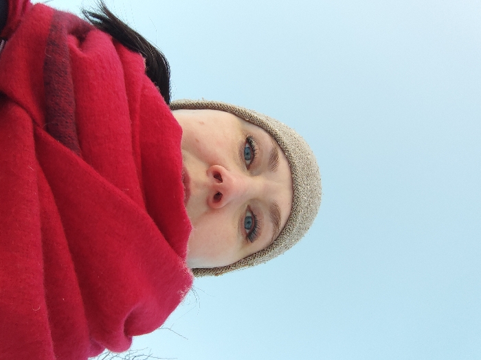 A low-angle close-up image captures an adult individual, reportedly located in the city of Chaikovsky, Russia; however, the precise location within the city is not visually discernible from the image. The subject's face is partially visible, from the upper cheeks and nose upwards, featuring blue eyes with dark eyelashes and light eyebrows. A light brown knitted beanie hat covers the head, with dark hair visible on the left side. A substantial red scarf, displaying a darker red horizontal stripe on its left fold, obscures the individual's mouth and lower face. The subject is looking directly into the camera. The background is a uniform, clear light blue sky, indicating an outdoor, daytime scene. No other environmental features or objects are clearly distinguishable.