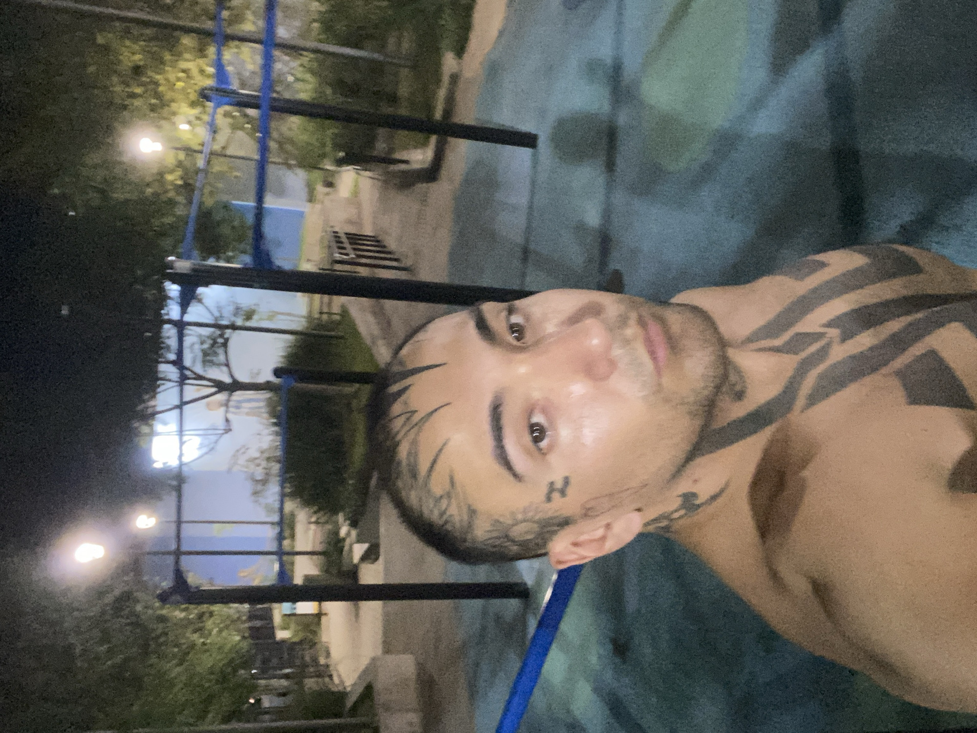 The image shows a man with tattoos on his face, neck, and shoulder, outdoors at night. Streetlights illuminate the scene. The man is near a metal jungle gym structure, likely a part of a public park or exercise area. The location is likely Buenos Aires, Argentina based on the metadata. The surface beneath the man and the gym appears to be painted or textured. The man's expression is neutral. He is topless.