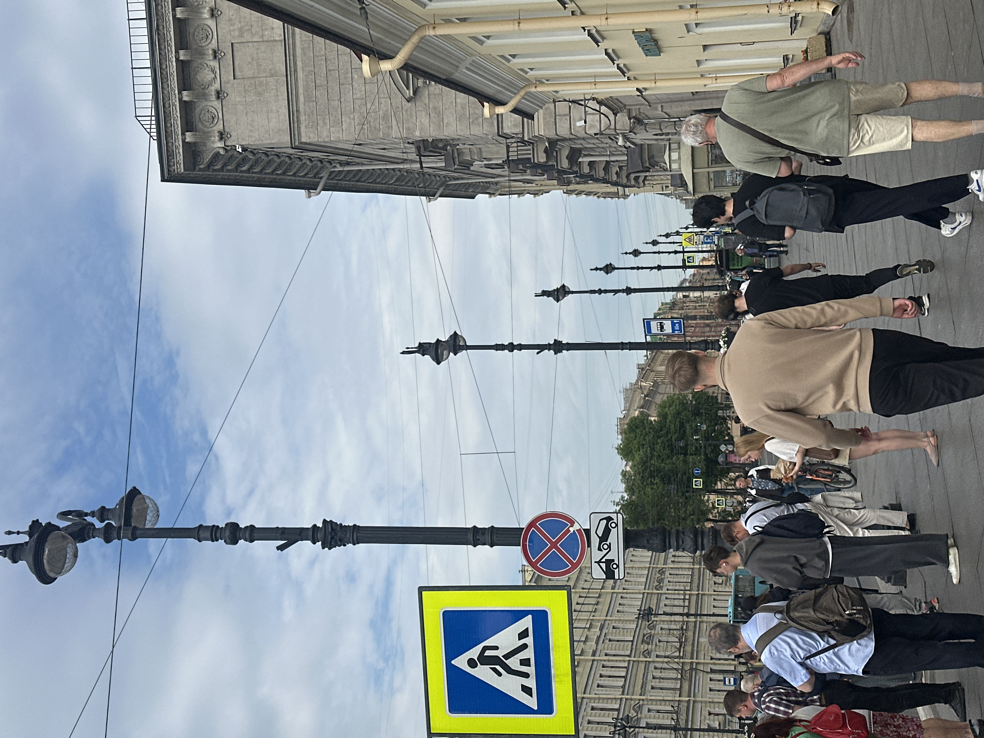 The image depicts a street scene in an urban environment. In the foreground, several individuals are walking along a sidewalk. A man in a tan sweatshirt and black pants walks away from the camera. To his left, a man in a grey sweatshirt and black pants, also with a backpack, walks in the same direction. Further to the left, a man in a green t-shirt and khaki shorts, carrying a messenger bag, walks away from the camera. Behind him, another man in a black t-shirt and black pants walks in the same direction. A woman in a white top and light-colored shorts stands to the right, holding a paper bag.
Several streetlights with multiple lamps are visible along the sidewalk, as are traffic and pedestrian signs. One sign displays a pedestrian crossing symbol within a blue square and yellow border. Another sign is circular with a red border and a blue background, featuring a red diagonal cross, indicating no stopping. Below this is a rectangular sign depicting a tow truck towing a car. Buildings line the right side of the street, with ornate architectural details. The sky is overcast with scattered clouds. In the background, more buildings and trees are visible, along with a light blue bus.