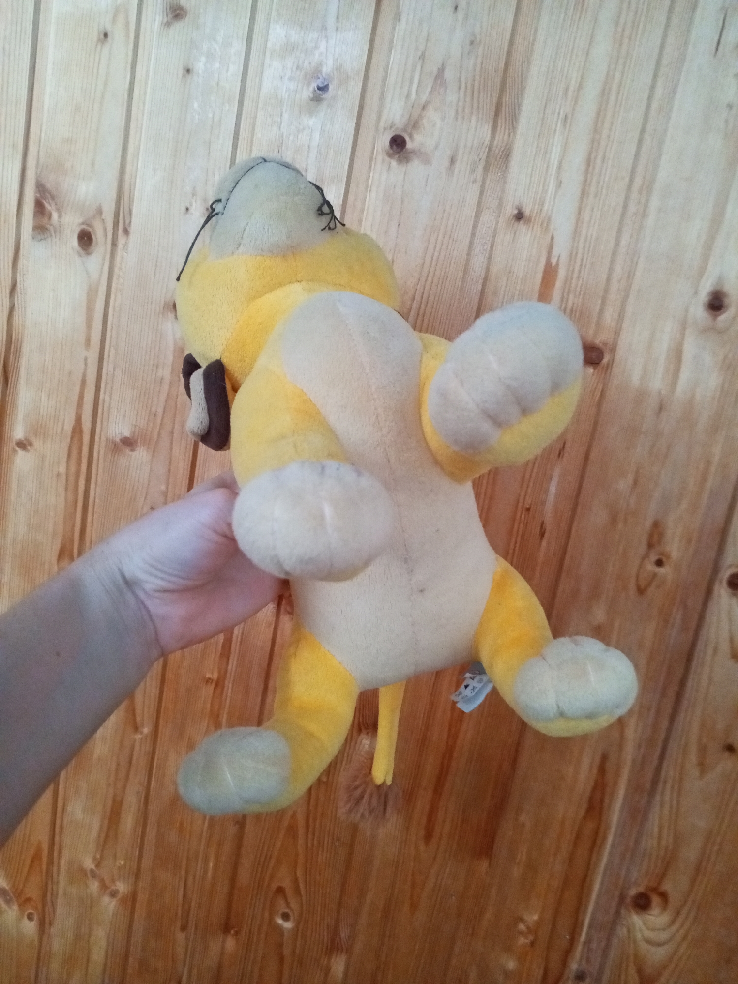 A yellow and grey plush toy, possibly a cat or dog, is held from the left side by a person's hand. The toy is positioned on its back with its legs in the air. The background consists of a wooden wall with vertical planks. The scene is indoors.