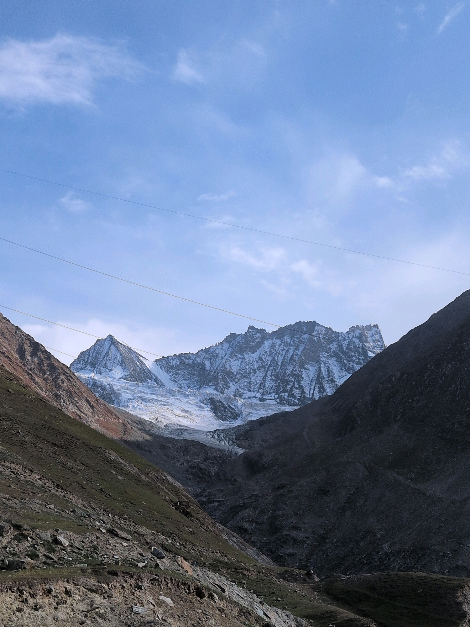 The image depicts a mountainous landscape, likely in a remote, high-altitude region. A prominent snow-capped mountain range dominates the background, with a glacier visible at its base. The surrounding slopes are rugged and rocky, with patches of sparse vegetation. The sky above is a pale blue with wispy clouds. Two thin power lines are stretched horizontally across the upper portion of the image, suggesting the presence of human infrastructure in this otherwise wild environment. The foreground shows a steep, rocky incline covered with low-lying greenery and loose stones. The lighting suggests it is either early morning or late afternoon, casting long shadows. The location is not within a city but rather in a natural, mountainous area.