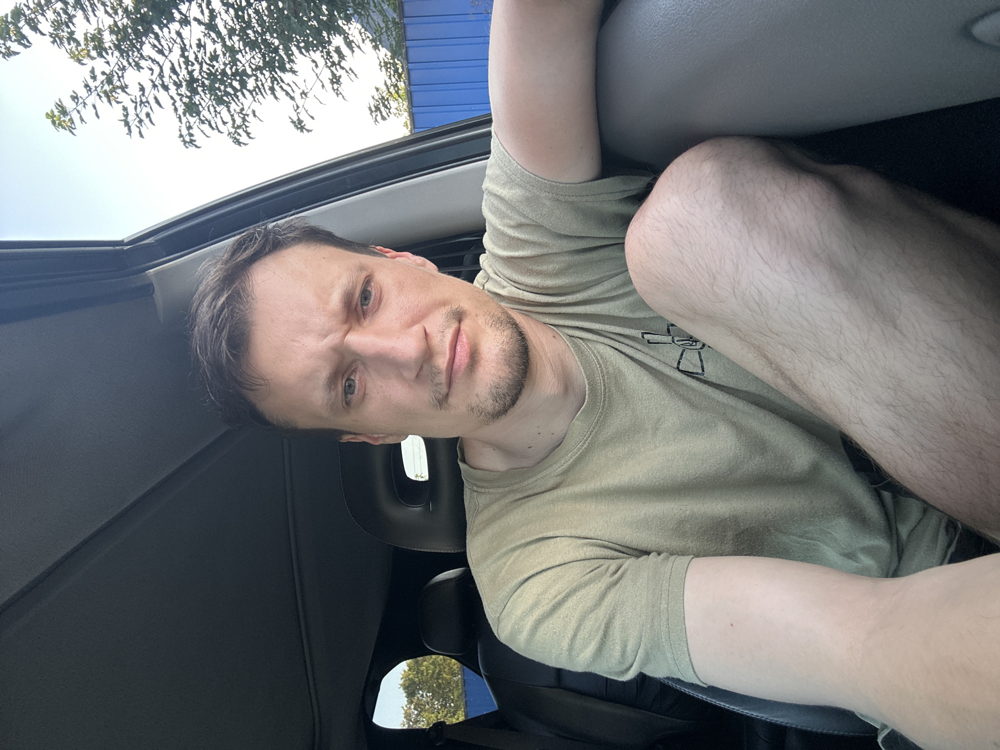A man is sitting in the driver's seat of a car. He is wearing a light green t-shirt. The car's interior is visible, including the headrest and the passenger seat. Outside the car, trees and a blue fence can be seen. The image appears to be taken in the city of Shatava, Ukraine.