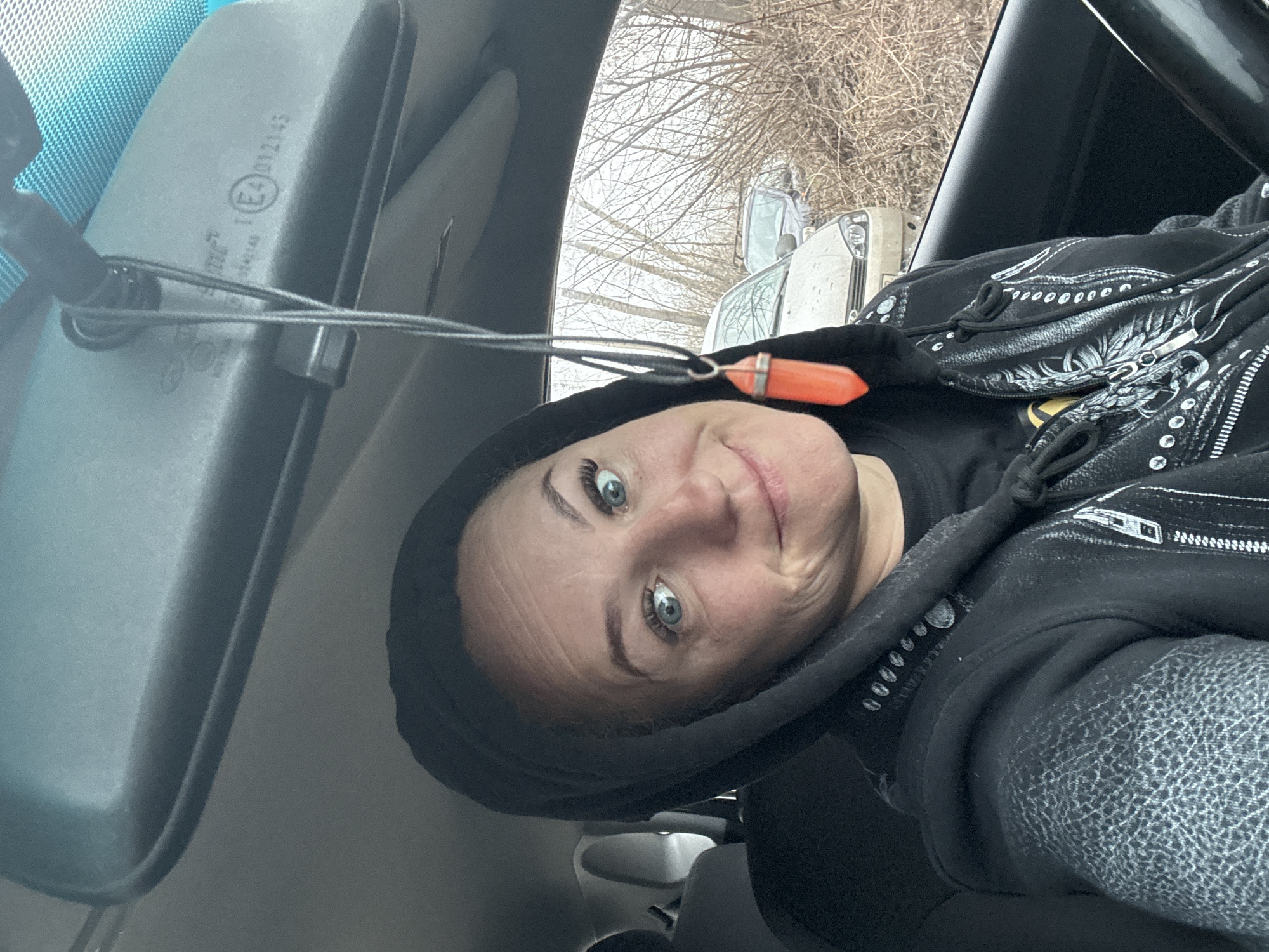 A selfie taken from inside a vehicle shows a woman with blue eyes wearing a black hoodie. The rear-view mirror is visible in the upper left. Outside the window, bare trees and a white vehicle are visible. Although the city is mentioned in the prompt, there is no visual indication to confirm the location of Chaikovsky, Russia. Therefore, I will exclude that from the description.