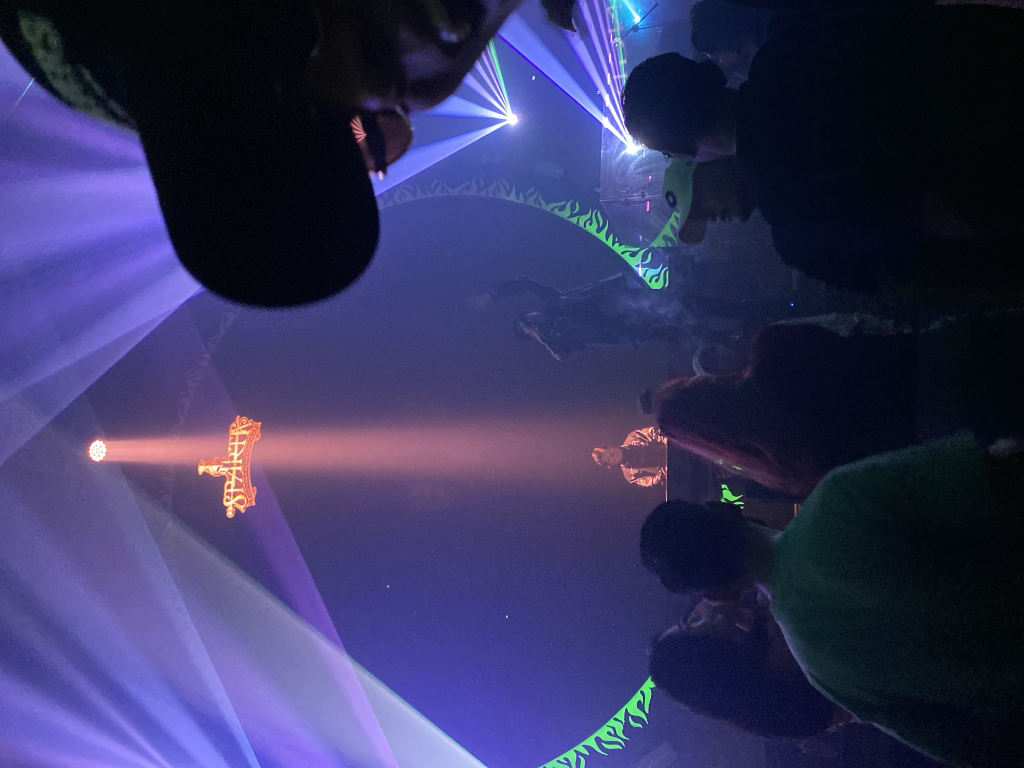 The image depicts an indoor event with a DJ booth at the center, illuminated by dramatic lighting. A DJ, wearing a jacket, is seen behind the booth, likely performing. Above the DJ, a red sign with the word "SPARTA" is prominently displayed, lit by a spotlight. The venue appears to be a club or concert hall, with a crowd of people in the foreground, silhouetted against the stage lighting. The atmosphere is dark, with vibrant purple and blue lights creating beams that fan out across the space. The overall scene suggests a lively nightlife setting in South Jakarta, Indonesia.
