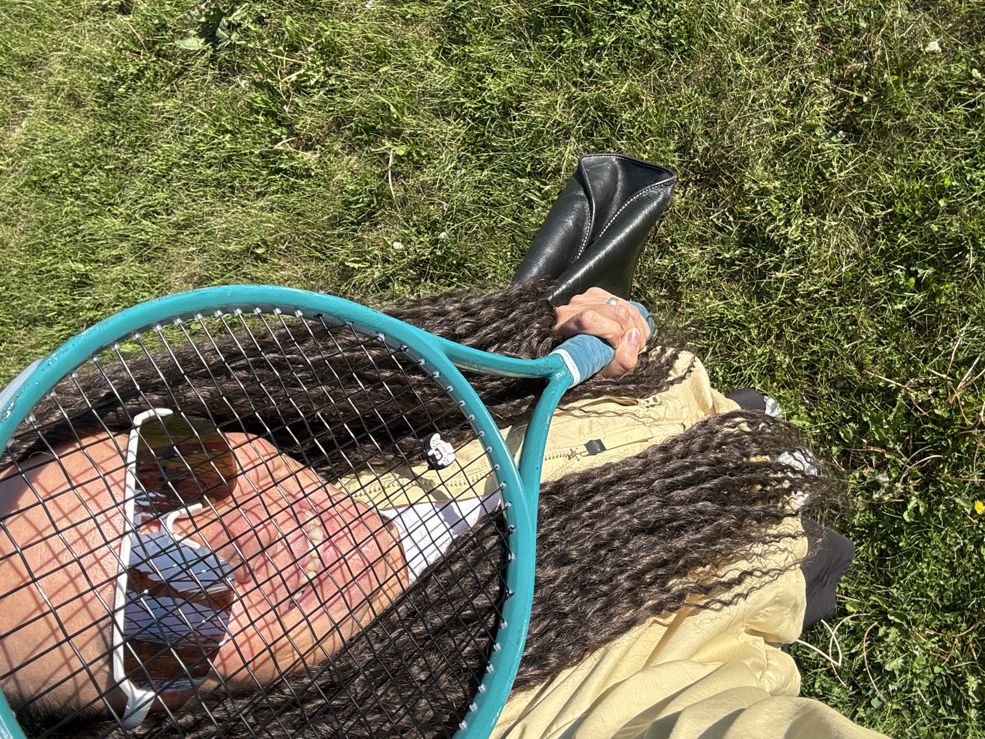 In Chaikovsky, Russia, a person is holding a teal tennis racket, the strings of which obscure their face. They are wearing white sunglasses and a light yellow collared shirt. Their long, dark dreadlocks extend over their shoulders and chest. In their left hand, they hold a black clutch bag. The person is standing on a grassy area under a clear, sunny sky.
