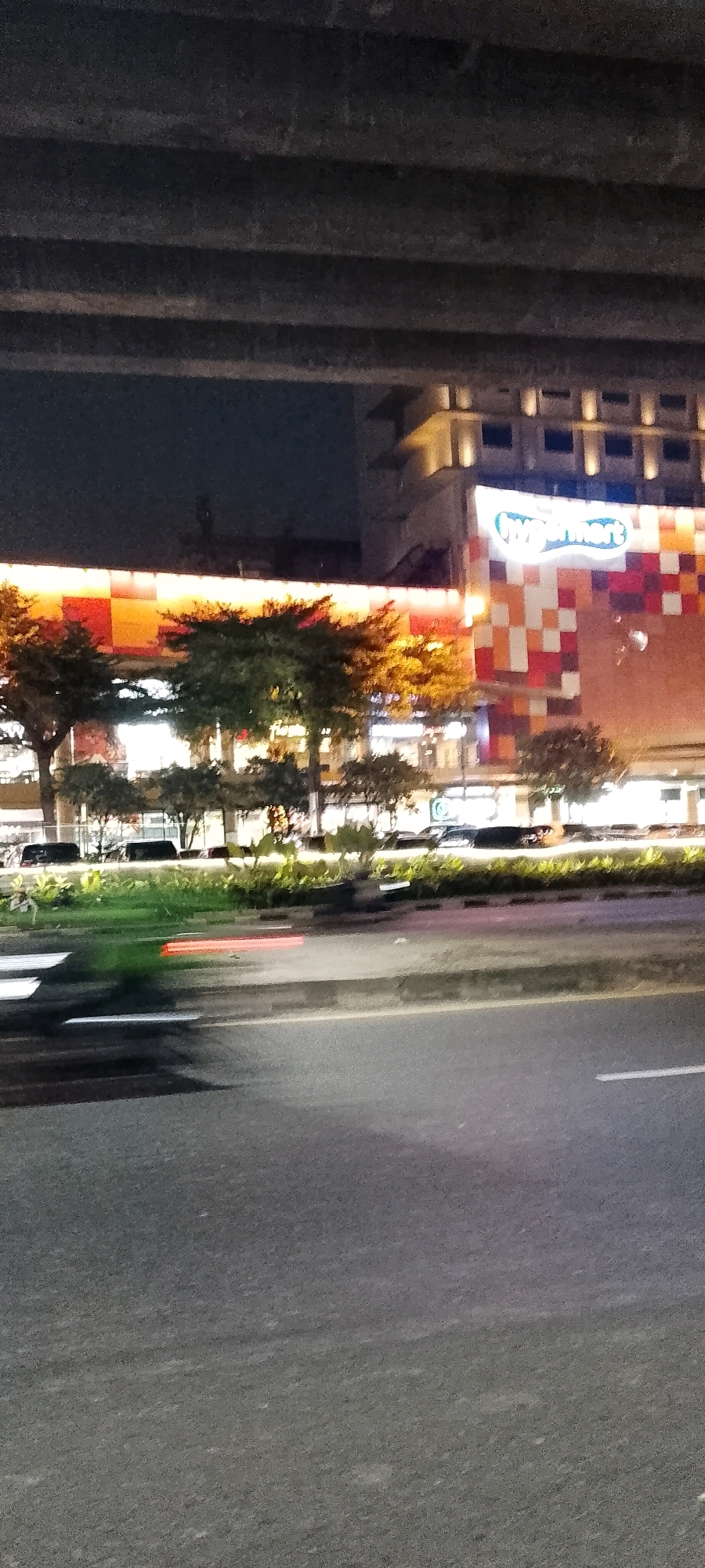 This is a night-time, slightly blurry, vertical, wide-angle shot of a city street in Bekasi, Indonesia. The foreground is dominated by a dark asphalt road, with visible white lane markings and a gray concrete curb. To the left of the road, a green median strip is partially visible with some red and white barriers, possibly a pedestrian crossing. In the mid-ground, trees with sparse foliage line the median strip. Behind the trees, a building facade with a colorful, geometric pattern of orange, red, and white squares is illuminated. A large, white, oval-shaped sign with the word "hypermart" in blue, glowing letters is prominently displayed on the building. The building itself appears to be a commercial establishment, possibly a supermarket. Above the building, a dark, elevated concrete structure, likely an overpass or bridge, casts shadows. The sky above is dark, indicating it is nighttime. There are no people clearly visible in the image, but several dark-colored vehicles appear to be parked or moving along the street in the mid-ground, beyond the median. The overall lighting is dim, with the building's lights and the headlights of vehicles providing illumination.