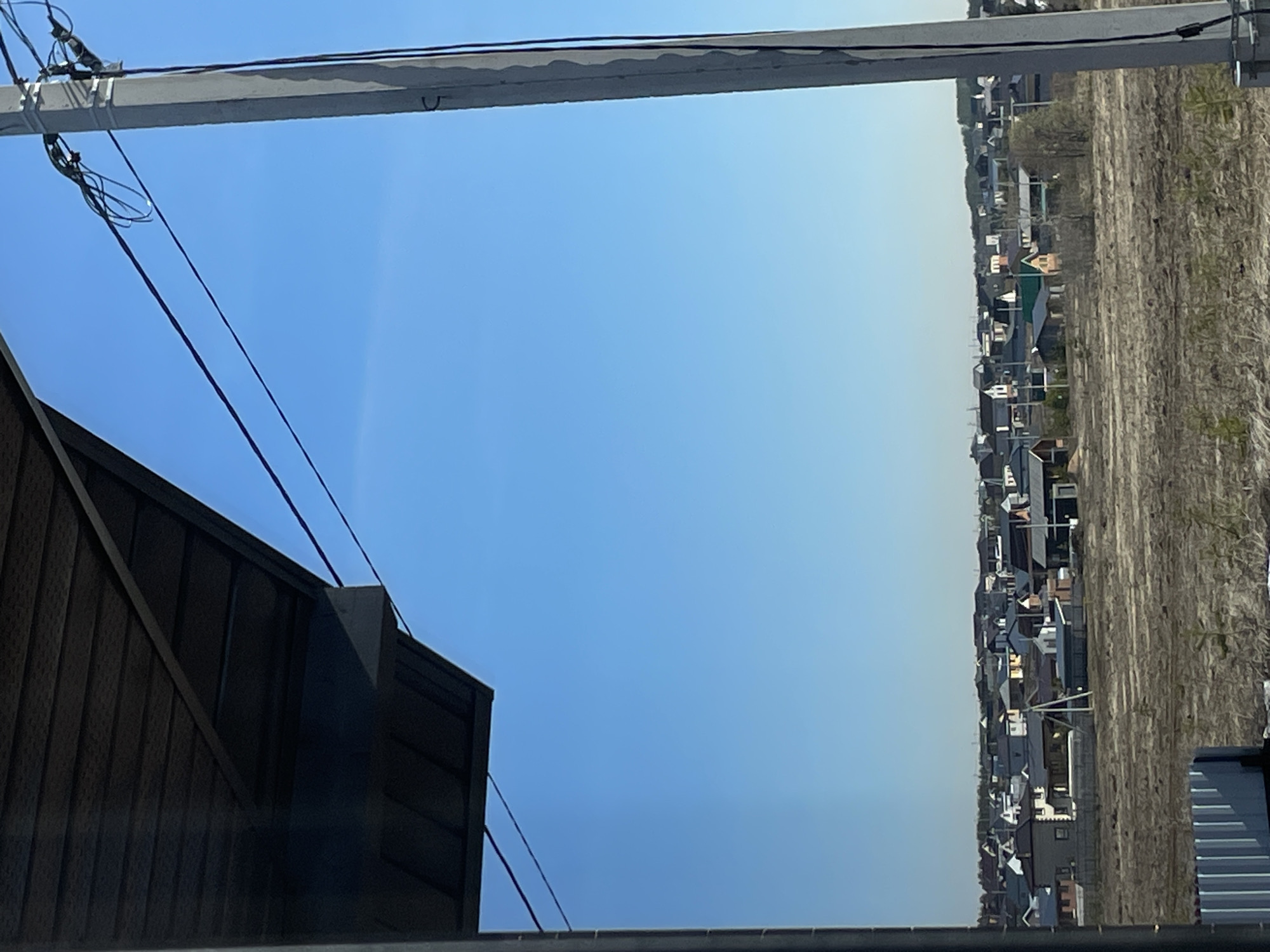 The image shows a view from an elevated position in what appears to be a suburban or rural area. The sky is clear blue, occupying the majority of the frame. A section of a dark-colored building is visible in the lower left corner, with vertical ridged siding and angular architectural features.
Electrical wires are strung across the upper left quadrant, attached to a concrete utility pole. In the distance, there's a row of closely packed houses and buildings. Beyond the buildings, there's a field or expanse of dry, possibly agricultural land.
The orientation of the scene suggests a location in a relatively flat area, given the long line of buildings and terrain. The proximity of the buildings and agricultural land suggests a transitional area on the edge of a town or city. However, the location as Подшивалово, Russia cannot be confirmed or disproven using only the visual information in the image.