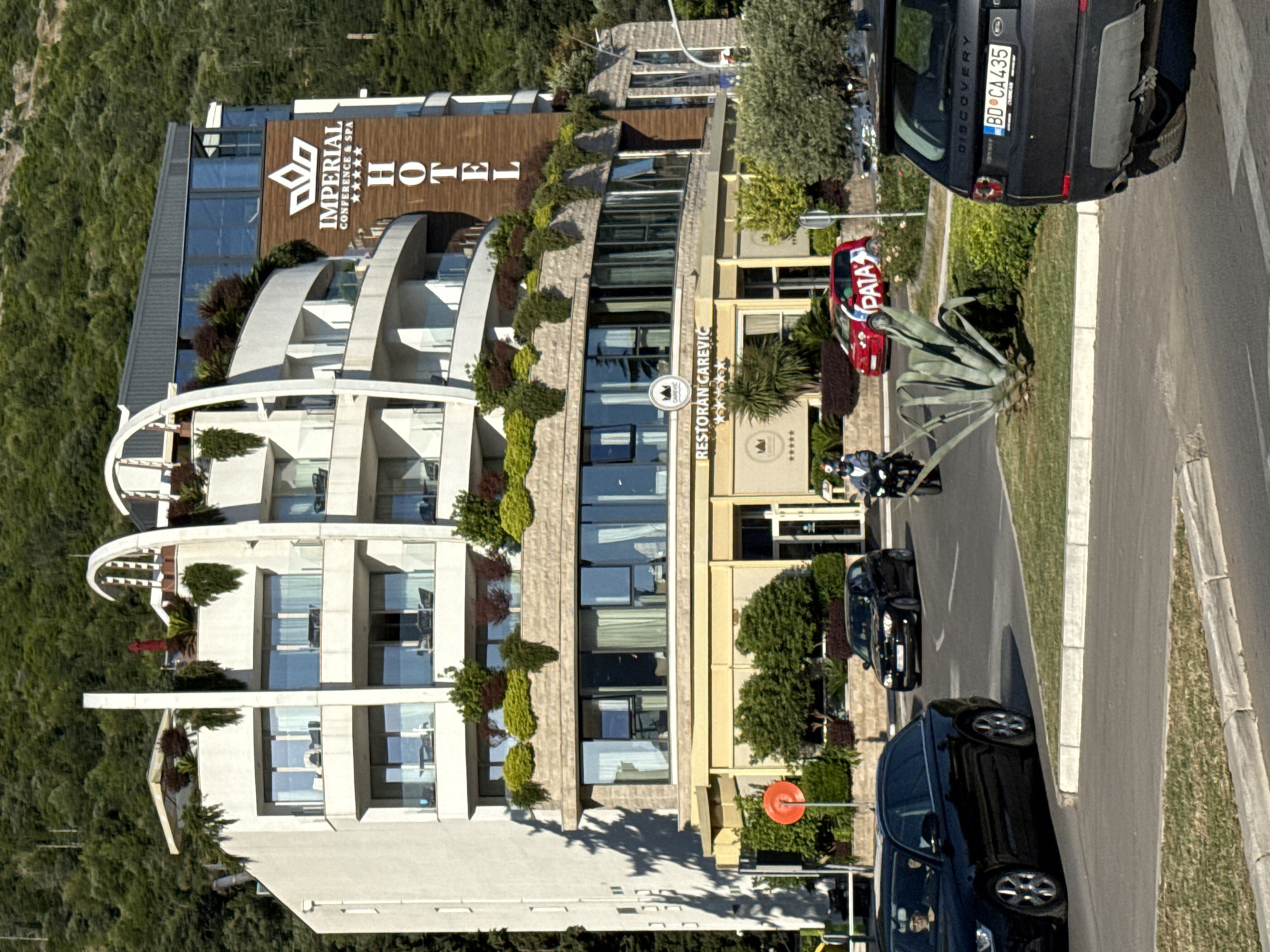 The image presents a daylight scene in Budva, Montenegro, centered on a multi-story building identified as the Imperial Conference & Spa Hotel and Restoran Carevic. The hotel, predominantly white with curved facades, features multiple balconies equipped with glass railings and adorned with extensive green landscaping, including shrubs and small trees. The "IMPERIAL CONFERENCE & SPA HOTEL" sign, indicating a five-star rating, is mounted vertically on a wooden panel on the building's right side. The ground floor, with a light yellow/beige facade and large windows, houses "RESTORAN CAREVIC," also displaying a five-star rating.
An asphalt road occupies the foreground, bordered by concrete curbs and grassy median strips. A black Audi station wagon, with a visible driver, is in the left lane, moving towards the viewer. Further down the road, a black BMW hatchback is present. A red Fiat 500, marked with "PATA" on its side, is positioned behind a large agave plant in a central median. In the right foreground, the rear of a dark grey Land Rover Discovery Sport is visible, bearing the Montenegrin license plate "BD CA435," confirming the location in Budva. A motorcyclist, wearing a helmet and a face mask, is on the left side of the road; another motorcyclist is partially visible on the far right. A red circular traffic sign is positioned on the left roadside. The background consists of a steep hillside covered with dense forest. Strong shadows on the left side of the hotel building indicate bright sunlight.