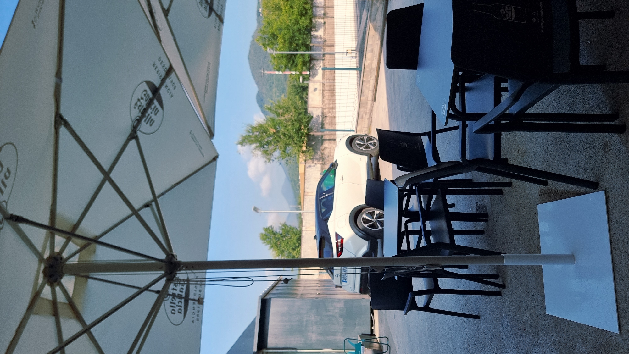 The image shows an outdoor seating area with several black tables and chairs, covered by a large white umbrella. The umbrella has a pattern of circular logos with text. In the background, a white car with a black roof is parked. Beyond the car, there is a fenced-off area and a building. Further in the background, trees and hills are visible under a clear blue sky. The ground is paved with concrete. The scene appears to be a commercial outdoor space, possibly a cafe or restaurant terrace. The location is identified as Olot, Spain.