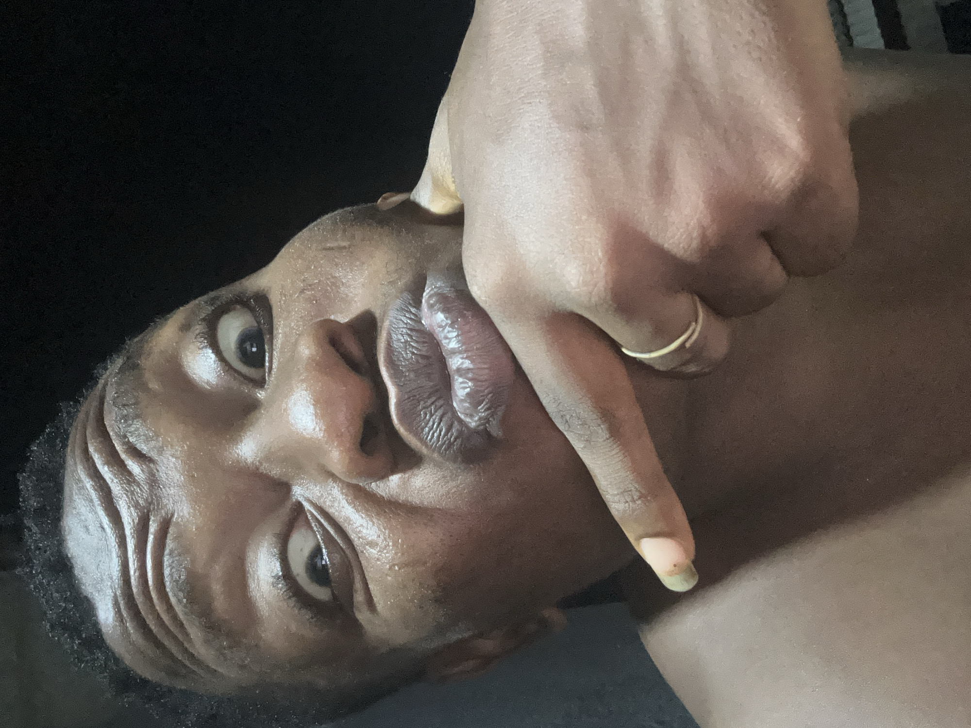A close-up portrait of an adult individual with dark skin, captured in a playful or exaggerated facial expression, understood to be in Ikorodu, Lagos State, Nigeria. The subject's forehead is deeply furrowed with multiple horizontal wrinkles, and the eyes are wide open, pupils visible, appearing to be looking slightly upwards and towards the viewer's left. The sclera exhibits a yellowish-reddish hue in some areas. The left cheek is visibly puffed out, and the lips are pursed into an exaggerated "kissing" or "duck" pose, showing textured surface details and a dark pink to purplish coloration. The skin on the face and visible upper torso appears shiny or moist, suggesting either perspiration or applied oil. A left hand is positioned prominently in the foreground, with the index finger extended and pointed towards the pursed lips. A thin, plain gold-colored band is visible on the ring finger of the left hand. The fingernail of the extended index finger has a pale tip with a noticeable greenish-dark discoloration beneath it. The background is uniformly dark and featureless, indicating a low-light environment, possibly indoors at night or in a dimly lit space. The lighting is direct and frontal, creating strong highlights on the subject's features.