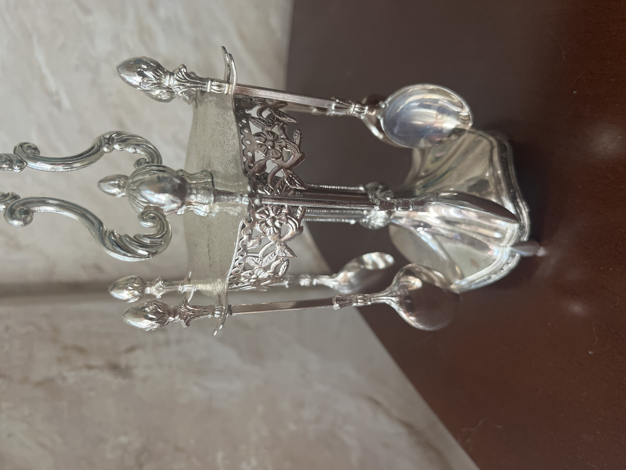 This is a close-up, eye-level, medium shot of a decorative silver-plated serving set with four small spoons suspended from a central stand. The stand is ornate, featuring a fluted base, a decorative filigree band with floral motifs, and a rounded top with scrolled elements. The spoons have small bowls and decorative handles. The set is placed on a dark brown, polished surface, likely a table. The background is a light-colored, textured wall that appears to be a marbled or faux-marble pattern. There are no people present, and no actions are being depicted. The image does not provide any information about the location in the city of Vladimirovka, Russia.