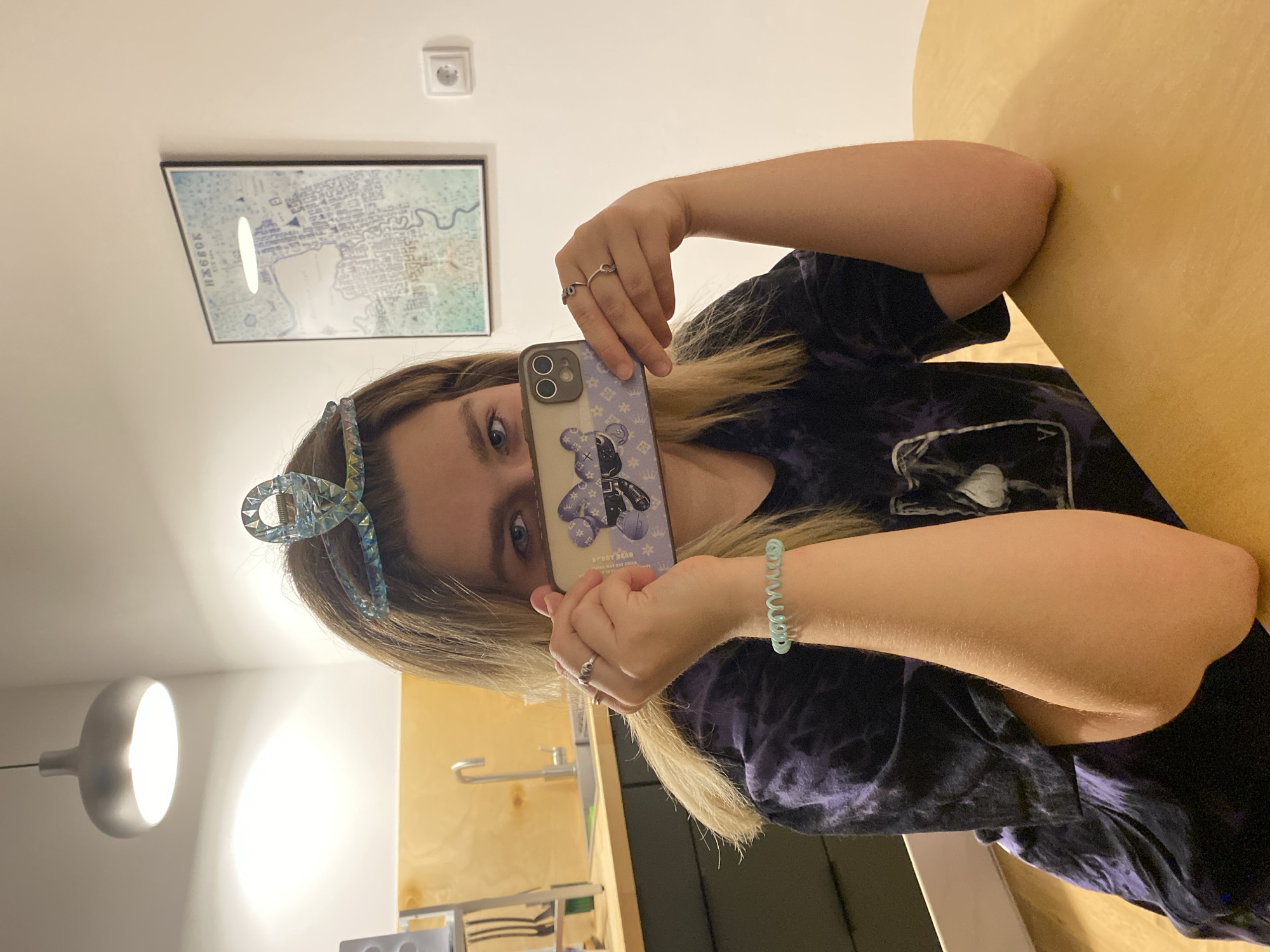 A young woman is taking a selfie indoors. She holds a phone with a decorative case featuring a cartoon bear in front of her face, obscuring most of it except for one visible eye. She has blonde hair with a jeweled blue hair clip. She is wearing a dark-colored t-shirt and a blue spiral bracelet. Two silver rings are visible on her fingers.
Behind her, a framed map hangs on the wall, along with an electrical outlet. The map features the plan of a city, specifically the city of Izhevsk, Russia. A modern-style pendant light illuminates part of the scene. A kitchen counter and sink are visible in the background, suggesting the location is possibly an apartment or house. A yellow surface, possibly a table or counter, is also visible.