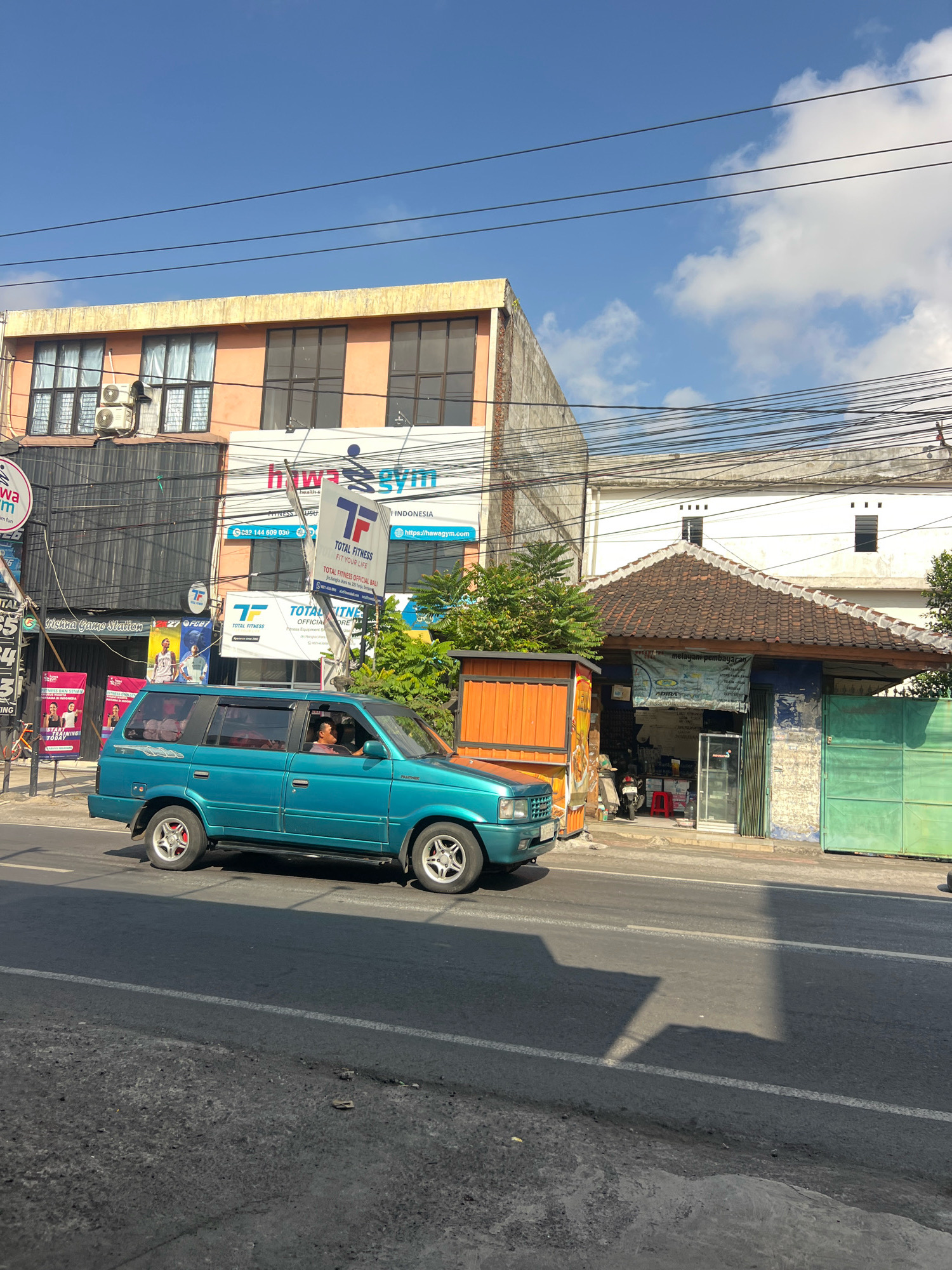 A street scene in Denpasar, Indonesia, captures a bright, sunny day. The foreground shows a section of asphalt road with a white lane marking. A turquoise SUV is driving on the road, its driver visible behind the wheel. The background features a row of buildings lining the street. One building has an orange facade with several windows. Another building displays a sign for "Hawa Gym" and "Total Fitness." Next to these is a smaller building with a tiled roof and an open storefront, displaying various items. A green gate is visible to the right of this storefront. Power lines crisscross the sky above the buildings. The overall impression is of a typical commercial street in a tropical city.