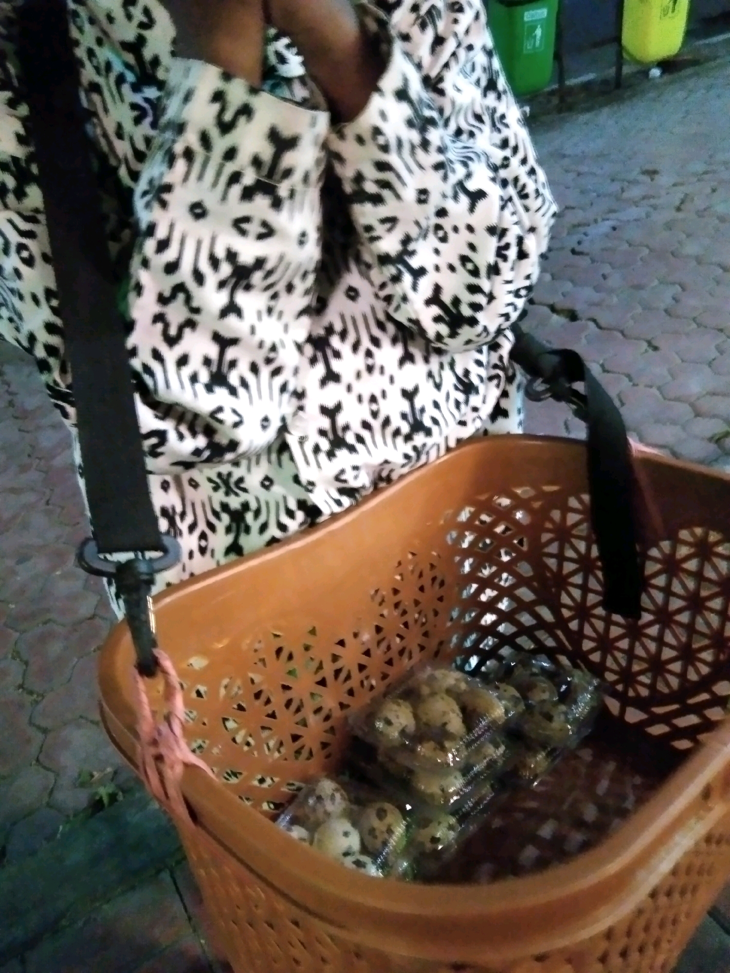 A person wearing a black and white patterned garment is seen from the waist down. They are carrying a brown plastic basket with a black strap attached to it via a pink string. Inside the basket, there are several plastic containers filled with quail eggs. The scene appears to be outdoors on a paved surface. In the background, two trash bins, one green and one yellow, are visible, suggesting an urban or public setting.