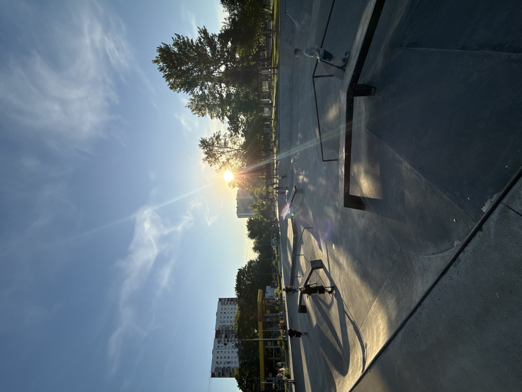 This image depicts a concrete skatepark at sunset within Khodynskoye Pole Park in Moscow, Russia. The sun is low on the right, casting long shadows across the skatepark features and generating lens flare. The sky is a light blue with wispy white clouds.
The skatepark occupies the foreground and mid-ground, featuring various concrete ramps, ledges, and rails. Approximately 10-15 individuals, predominantly children and young adults, are present within and around the skatepark. Several individuals are on scooters, including a boy in a green shirt and dark shorts on the right side, balancing on a rail, and another in a black shirt and dark shorts in the mid-ground left. Others are walking or standing.
In the mid-ground left, behind the skatepark, a yellow-and-red branded "ТАТНЕФТЬ" (Tatneft) gas station is visible, with multiple vehicles and people present. Directly behind the gas station, a large, multi-story residential panel building, predominantly white with red accents, rises against the sky. Further right in the background, a line of mature green trees partially obscures additional residential buildings and park elements, with another multi-story building visible beyond the tree line on the far right. The overall scene suggests a late afternoon or early evening recreational environment.