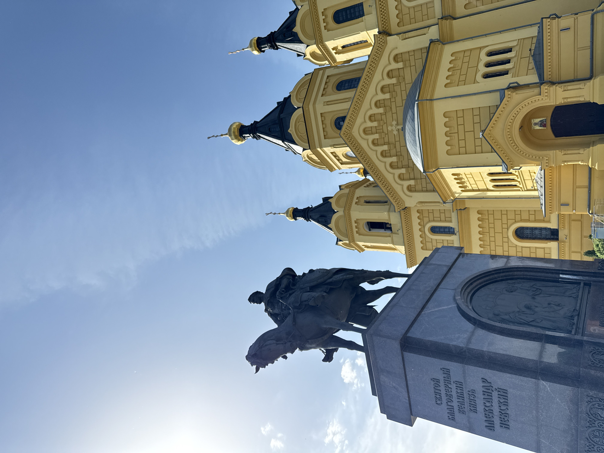 The image shows a view of Alexander Nevsky Cathedral and the Alexander Nevsky Monument in Nizhny Novgorod, Russia. The cathedral is a large, yellow building with multiple towers topped with black domes and gold spires. The monument depicts a bronze statue of Alexander Nevsky on horseback atop a dark gray granite pedestal. The inscription on the pedestal is written in Russian. The background is a clear blue sky.