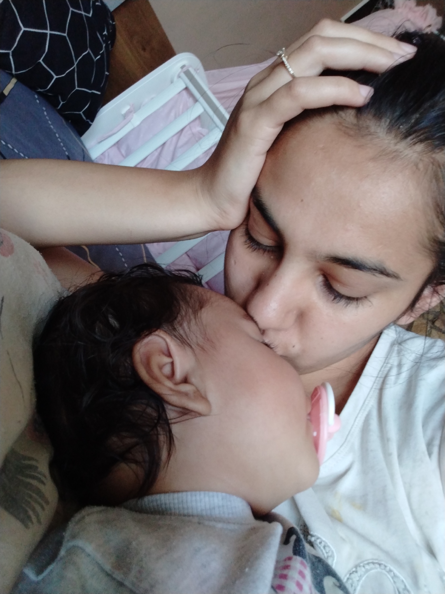 A close-up shot depicts a woman kissing a baby on the cheek. The woman has dark hair pulled back, dark eyelashes, and wears a ring on her right ring finger. She is wearing a white t-shirt. The baby is lying down with a pacifier in their mouth, and has dark curly hair. A patterned blanket and baby crib are visible in the background. Location: Unknown, Unknown.