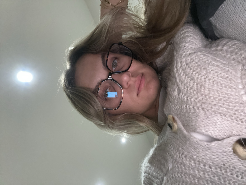 A woman wearing oversized square glasses looks at the camera. She has light brown hair that is parted to one side. She's wearing a fluffy cream-colored cardigan with large buttons. The background is a plain wall with several bright light reflections. A small portion of a dark gray pillow is visible behind her head.